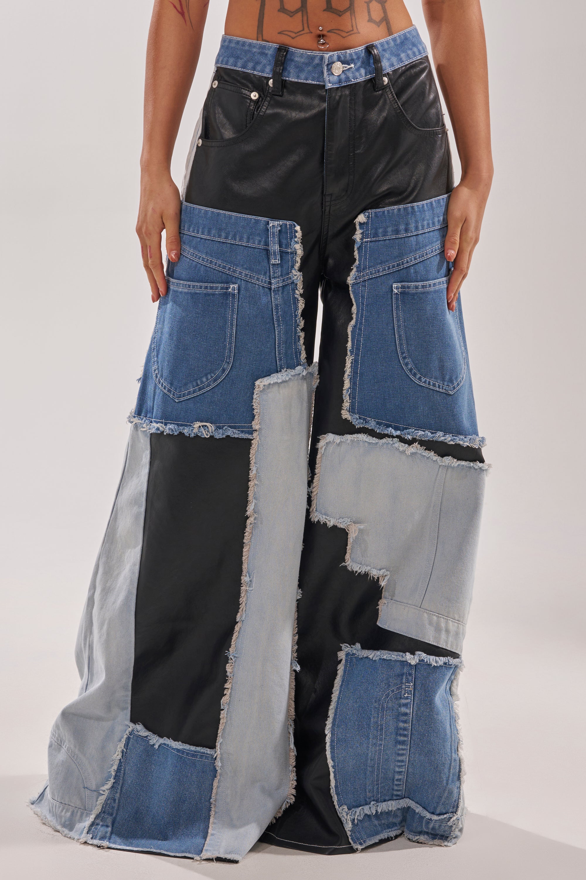 Someone shows off street style in the TOO MUCH GOING ON RELAXED DENIM, featuring wide-leg patchwork design, black faux leather accents, frayed edges, and oversized front pockets. Their exposed midriff and tattoo complete the edgy vibe. 4