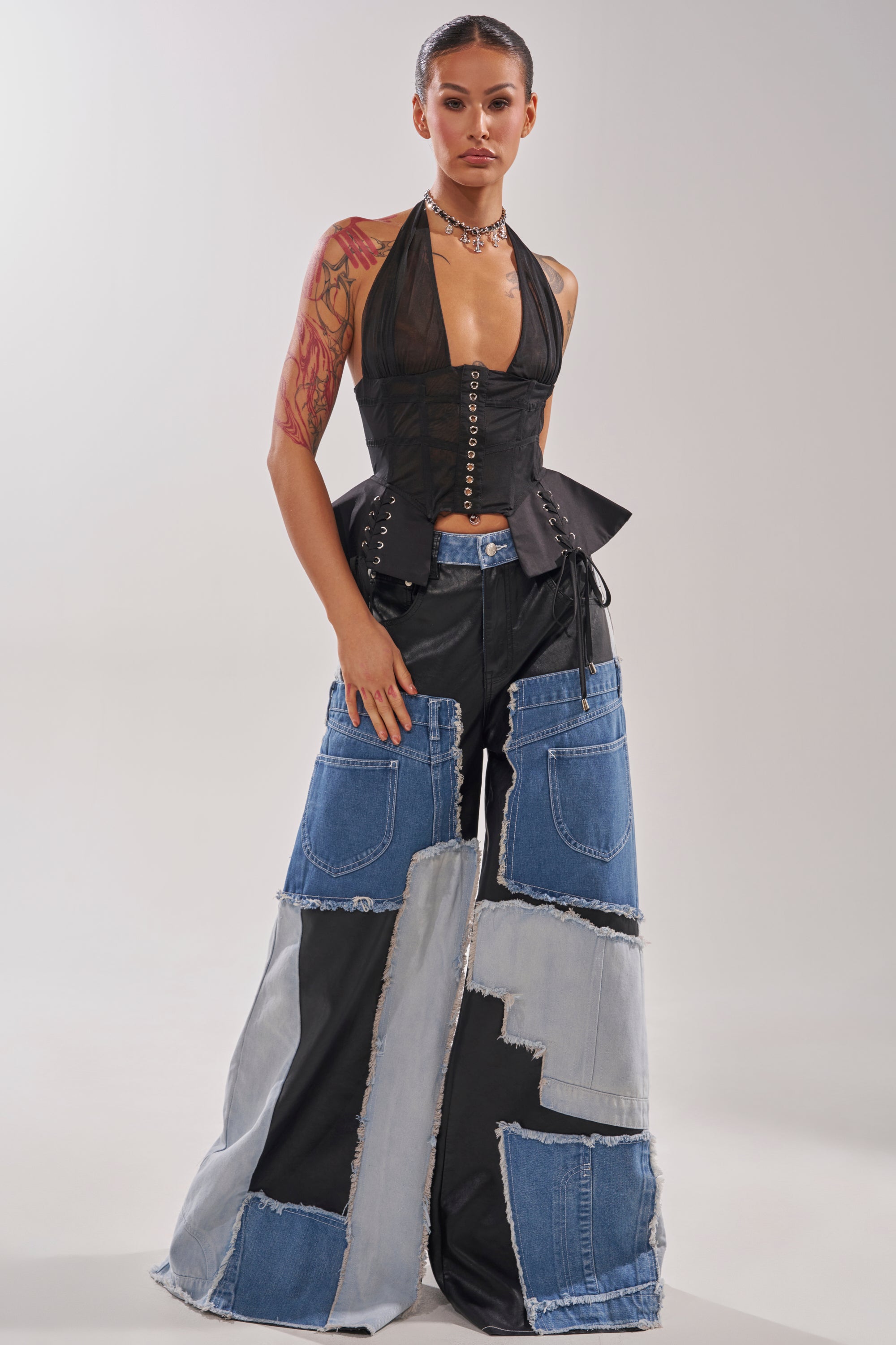 A woman exudes street style in a black lace-up halter top and the TOO MUCH GOING ON RELAXED DENIM pants. With a visible arm tattoo, sleek hair, and choker necklace, she poses confidently against a plain light background. 8