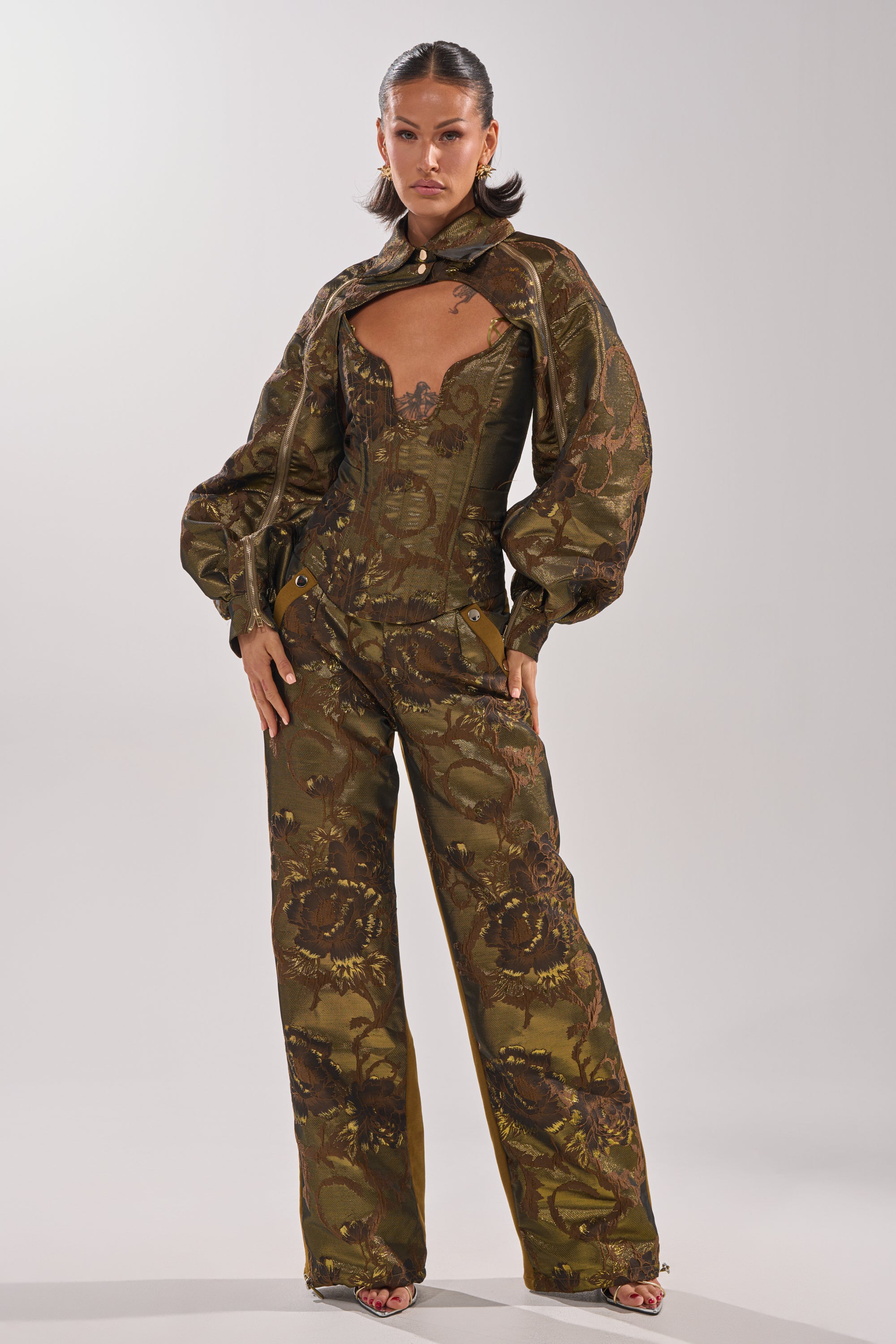 A woman poses confidently in the SHIGO BROCADE BOLERO, a metallic cropped jacket styled over a brown and olive green brocade jumpsuit with puffed sleeves, a structured corset bodice, and wide-leg pants against a plain light background. 11