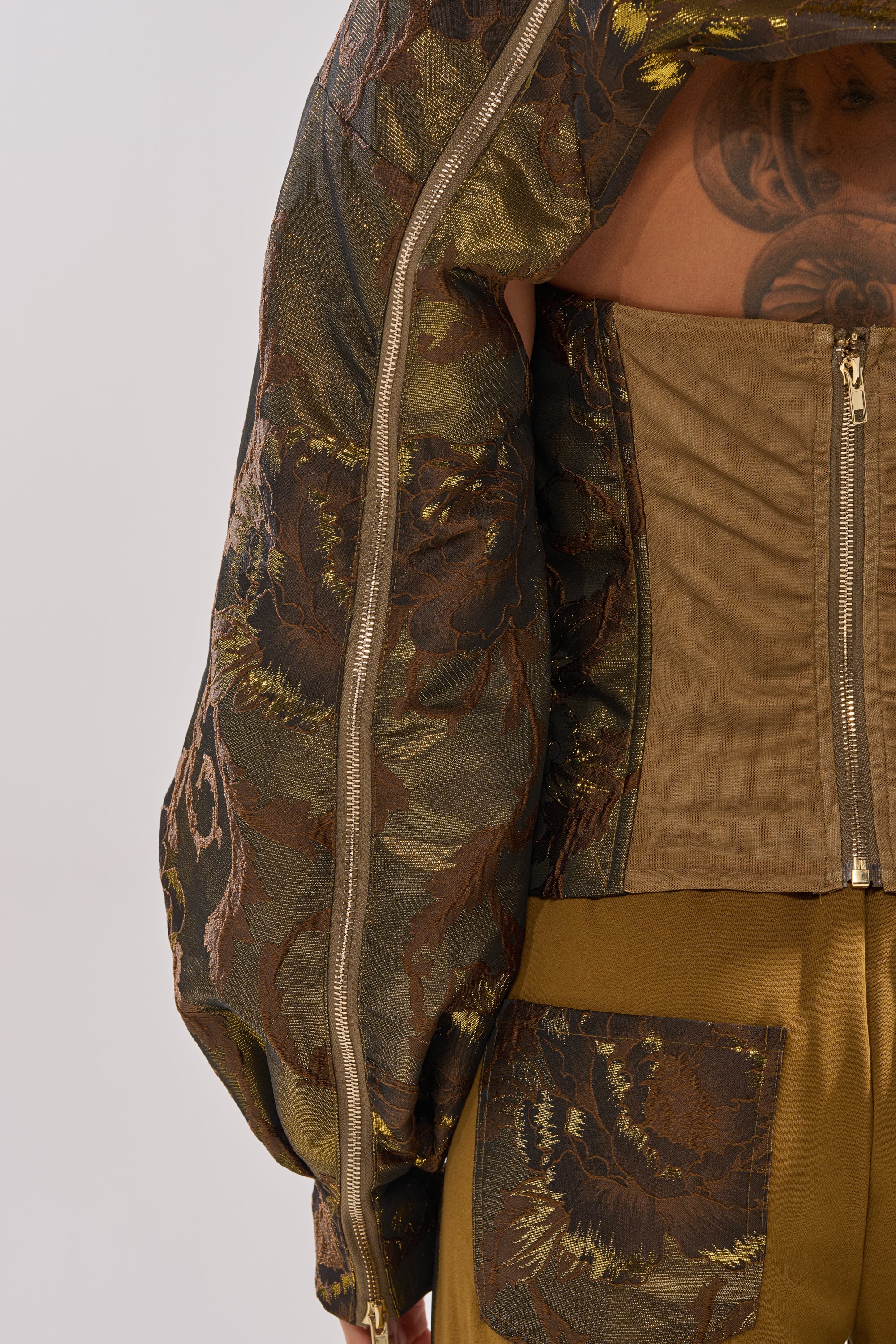 A close-up of a person wearing the SHIGO BROCADE BOLERO with textured floral patterns and zippers, styled with a structured tan corset, gold floral-pocket pants, and an upper back tattoo visible. 13