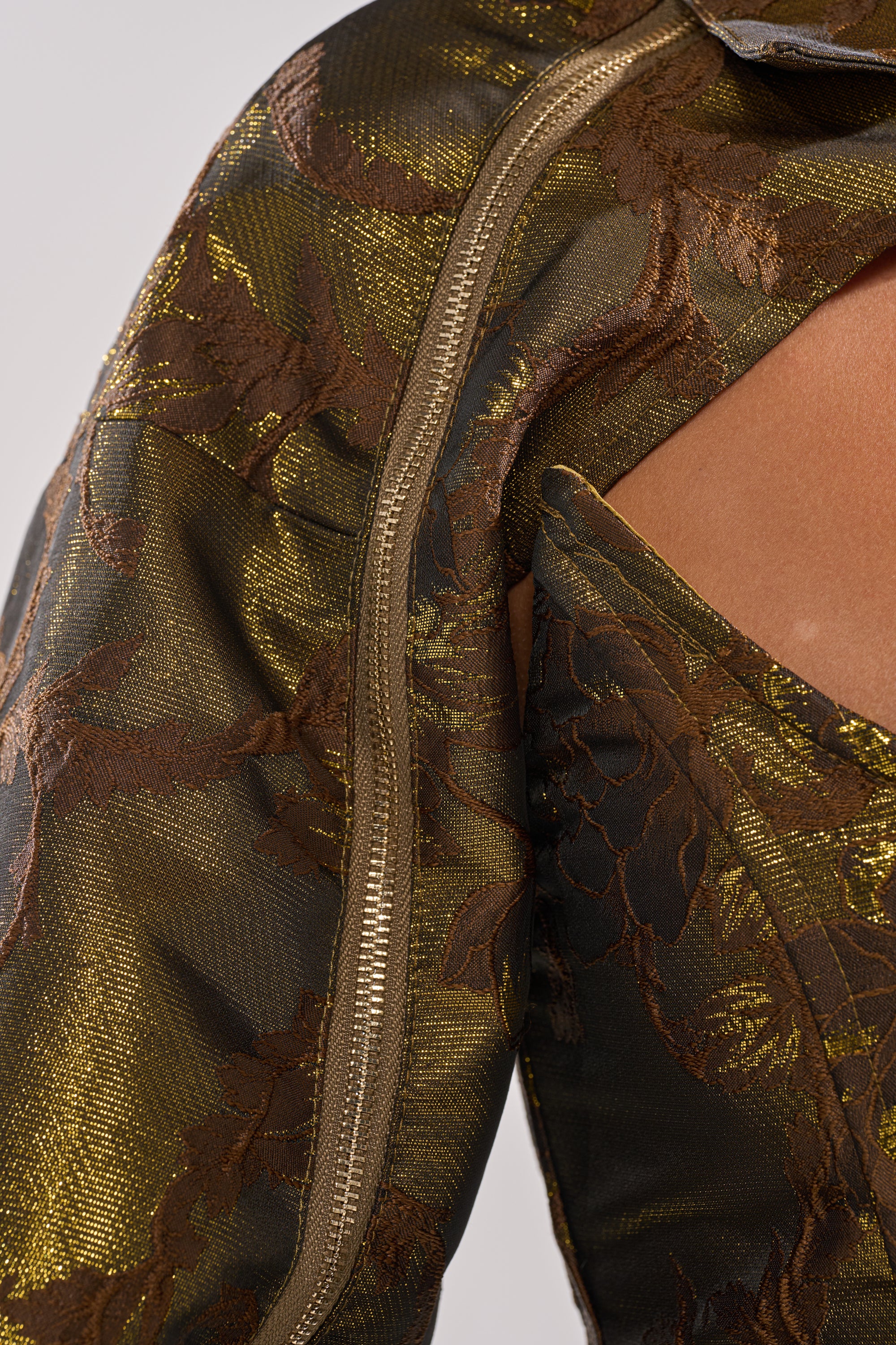 Close-up of the SHIGO BROCADE BOLERO, a cropped metallic gold and brown jacket with a floral jacquard pattern, zipper, and cut-out detail that reveals skin. The shiny texture enhances its sophisticated look. 14