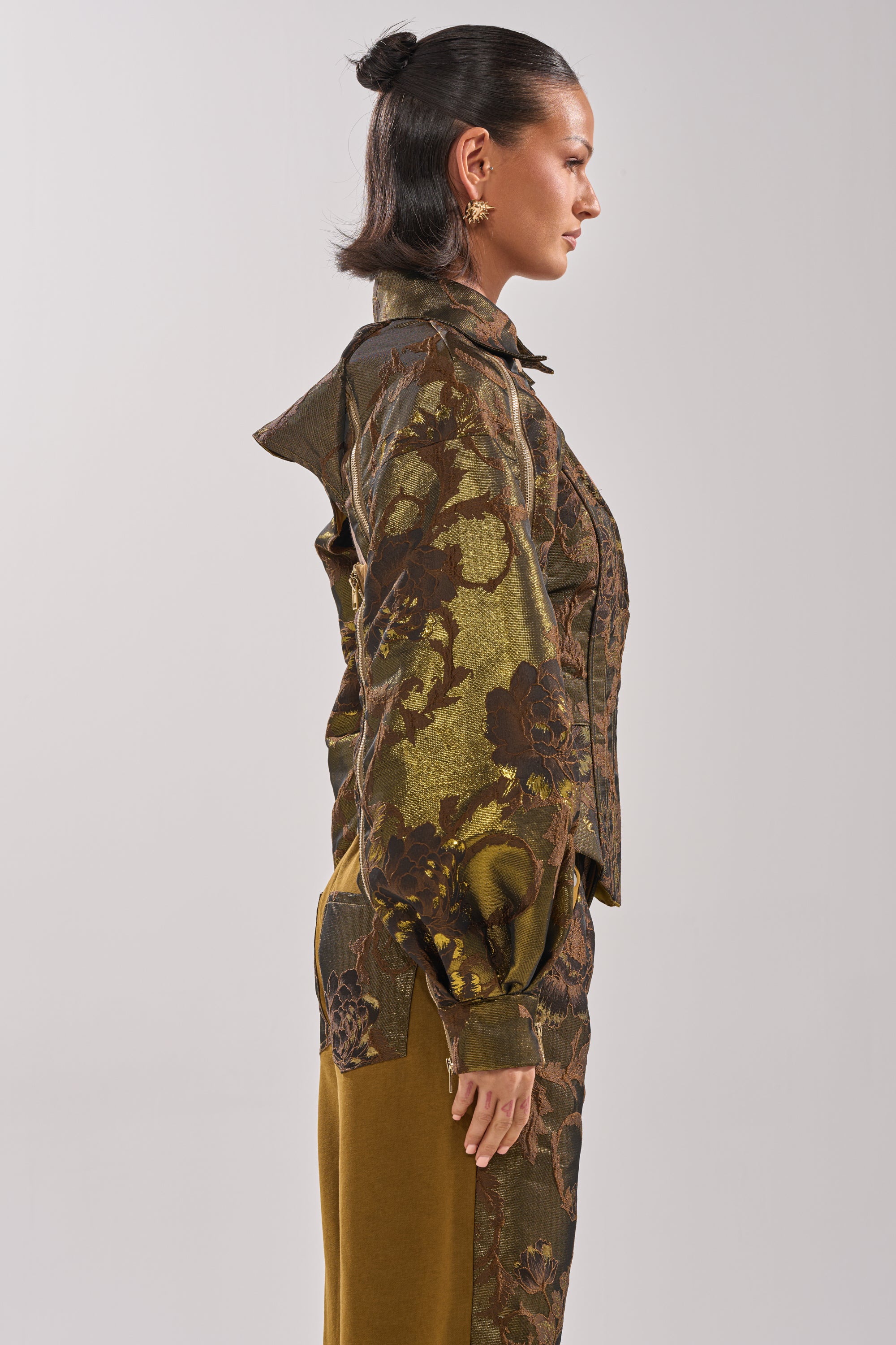 A woman in profile wears the SHIGO BROCADE BOLERO, showcasing jacquard fabric with green and brown florals and a subtle metallic sheen. Her hair is styled in a bun, gold earrings complete her look, and she poses against a plain light background. 4