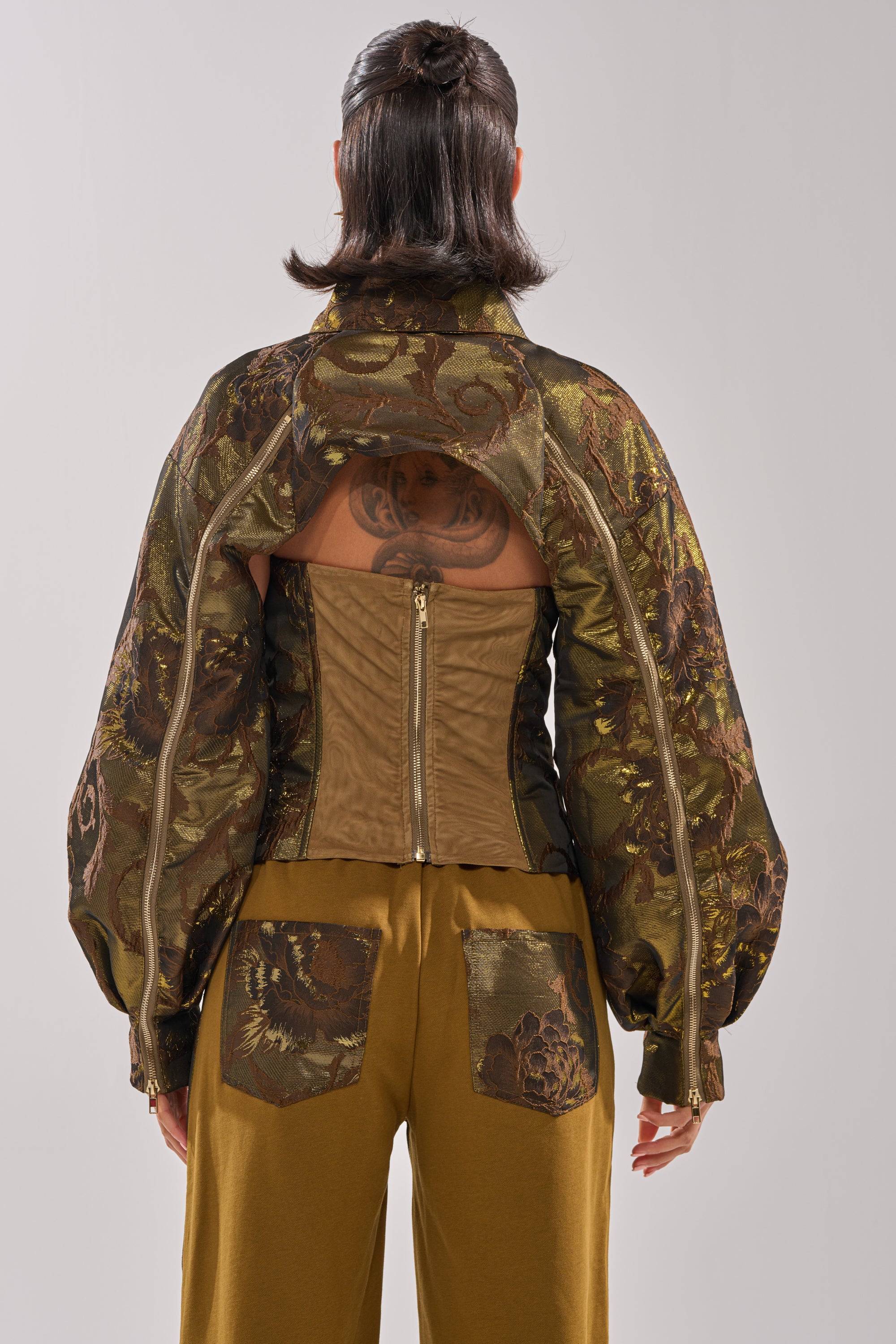 A person stands facing away, wearing the SHIGO BROCADE BOLERO with wide sleeves, unzipped to reveal a tan corset. They have mustard yellow patchwork pants and visible back tattoos. 5