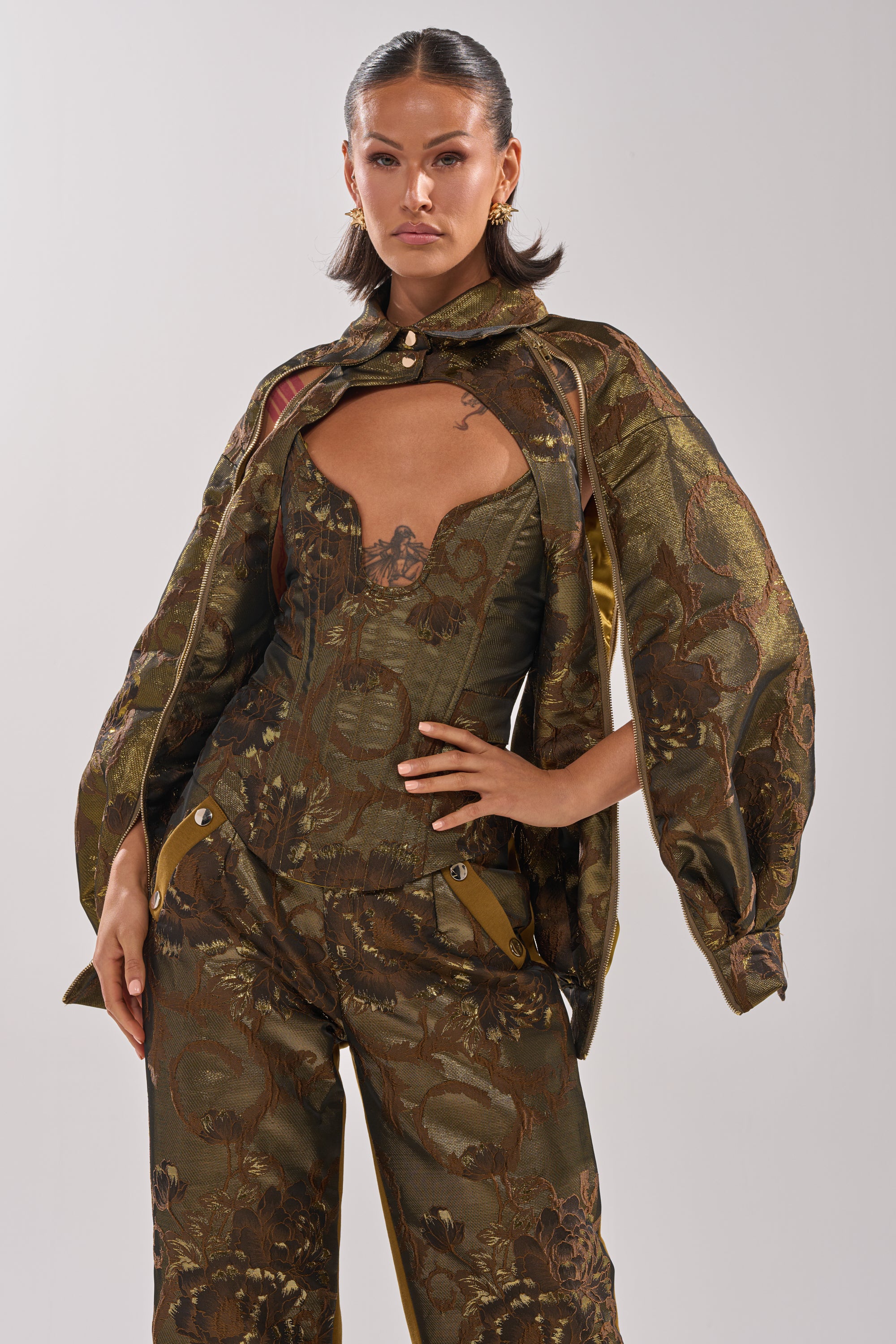 A woman with slicked-back hair and gold earrings confidently models the SHIGO BROCADE BOLERO in olive green, featuring a dramatic cape-style top, fitted corset bodice, and wide-leg pants against a plain background. 6