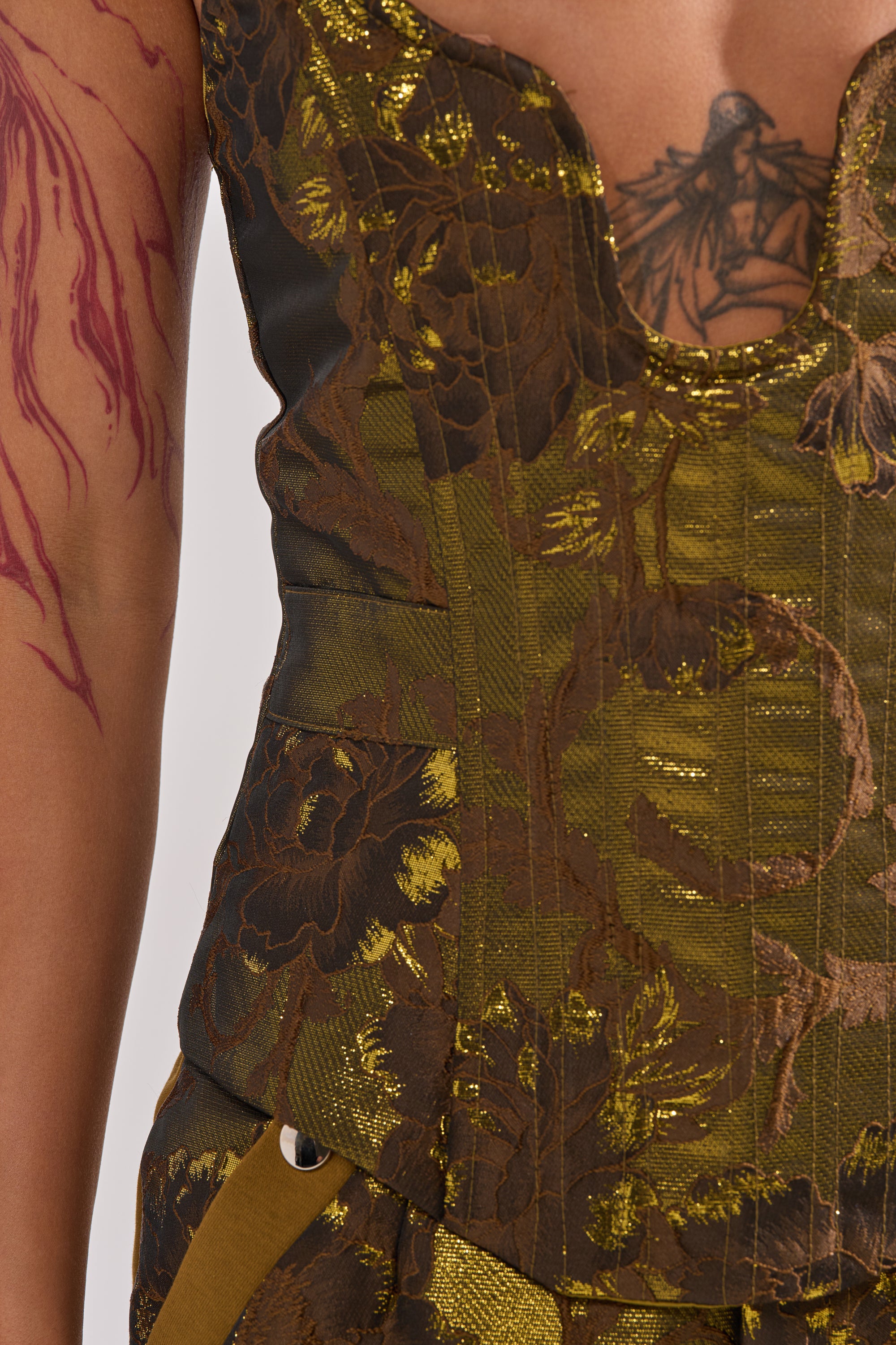 A close-up of a person wearing the SHIGO BROCADE CORSET in gold and brown jacquard. A detailed chest tattoo and red scar-like body art on their arm are visible. The background is plain white.