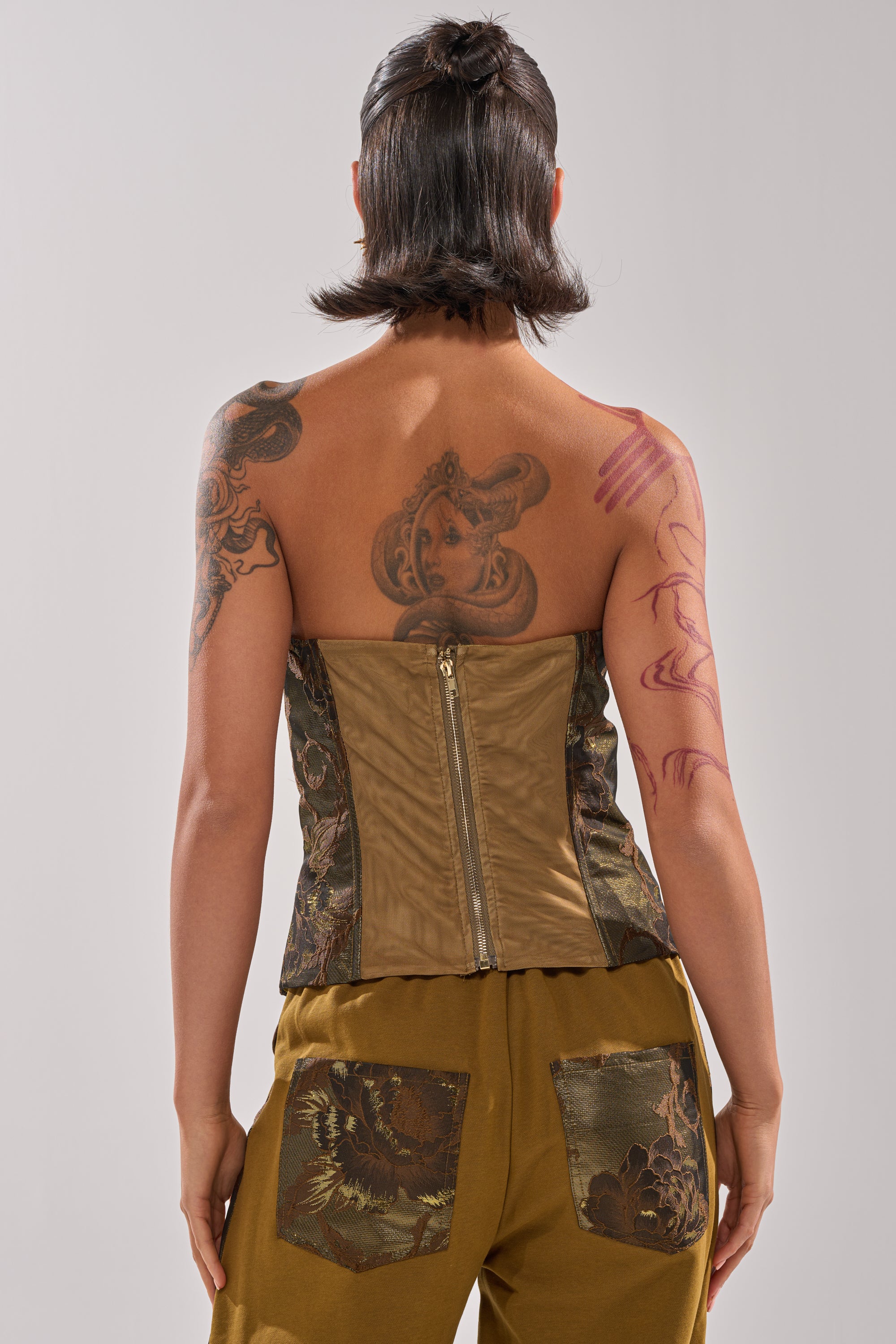 A person with short dark hair and visible tattoos wears the SHIGO BROCADE CORSET, a strapless zip-up top, paired with mustard pants featuring floral jacquard pockets, standing with their back to the camera.