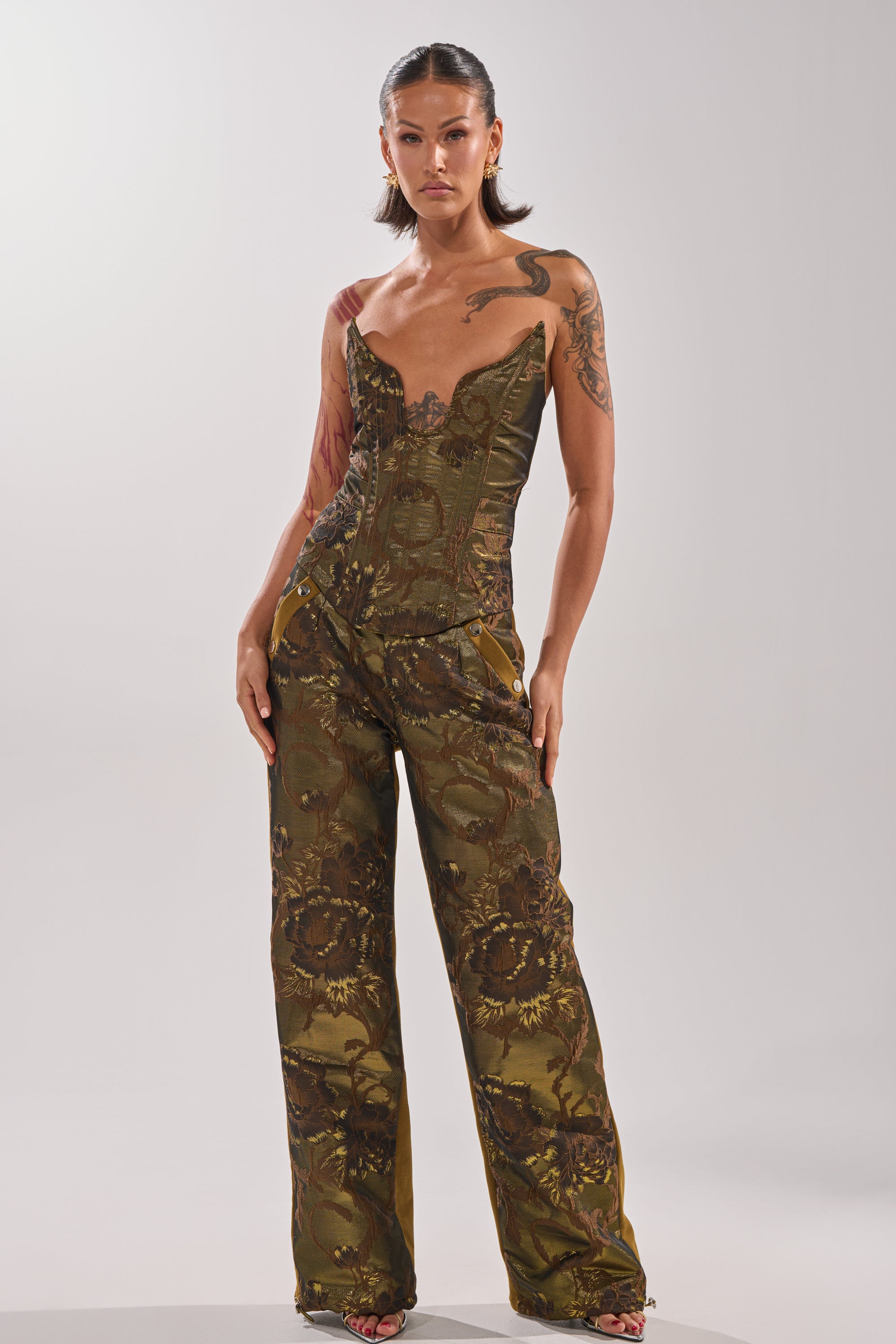 A woman with long hair pulled back and visible tattoos stands confidently in the SHIGO BROCADE CORSET, an olive-green, strapless jacquard top paired with matching floral wide-leg pants, set against a plain light background.