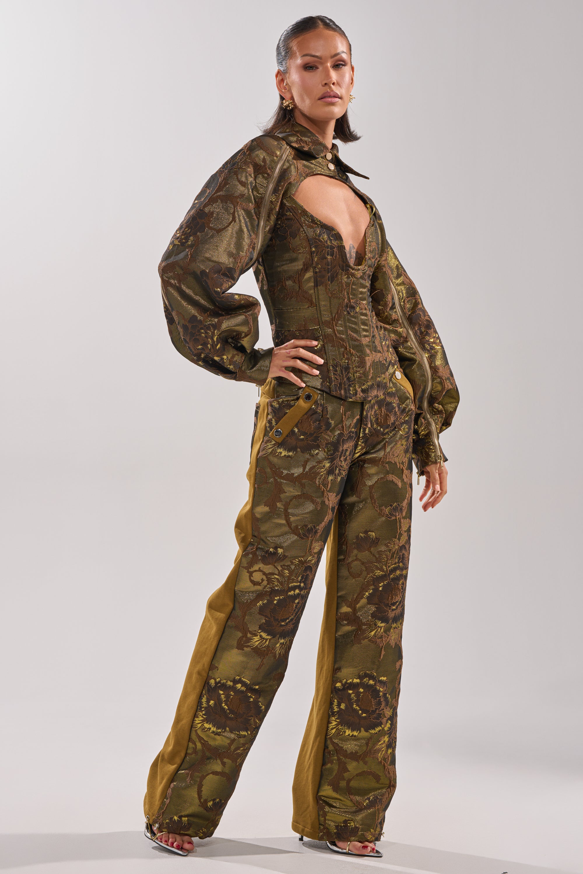 A woman stands confidently in the SHIGO BROCADE CORSET, a jacquard puff-sleeve top with a deep neckline and brown-green floral pattern, paired with matching wide-legged pants featuring bold yellow side panels.