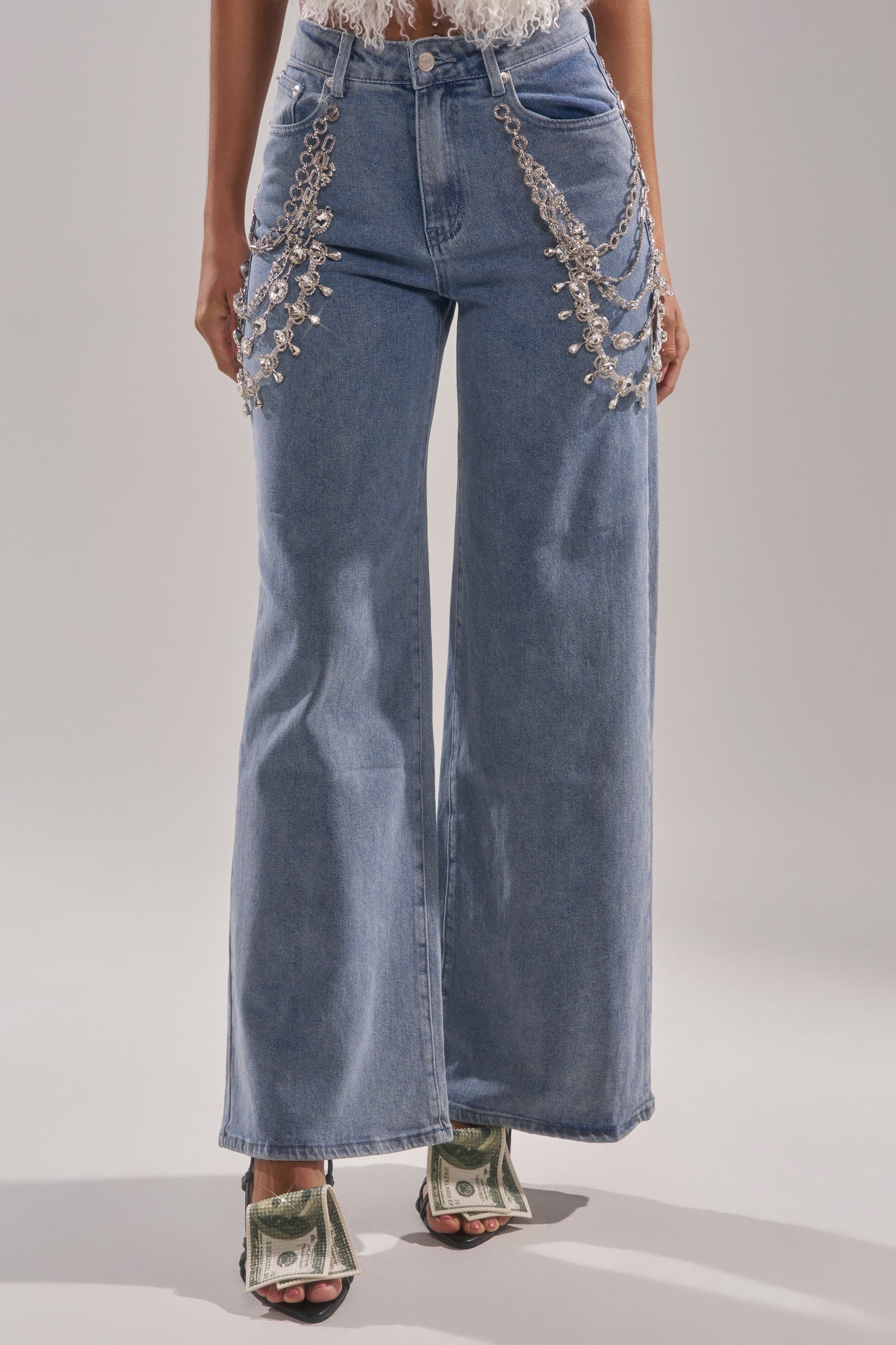A person stands on light gray ground wearing the DIAMOND LOVER RELAXED DENIM WITH RHINESTONE, showcasing wide-leg jeans with rhinestone embellishments. Their lower body is visible, paired with open-toe heels wrapped in U.S. dollar bills. 0