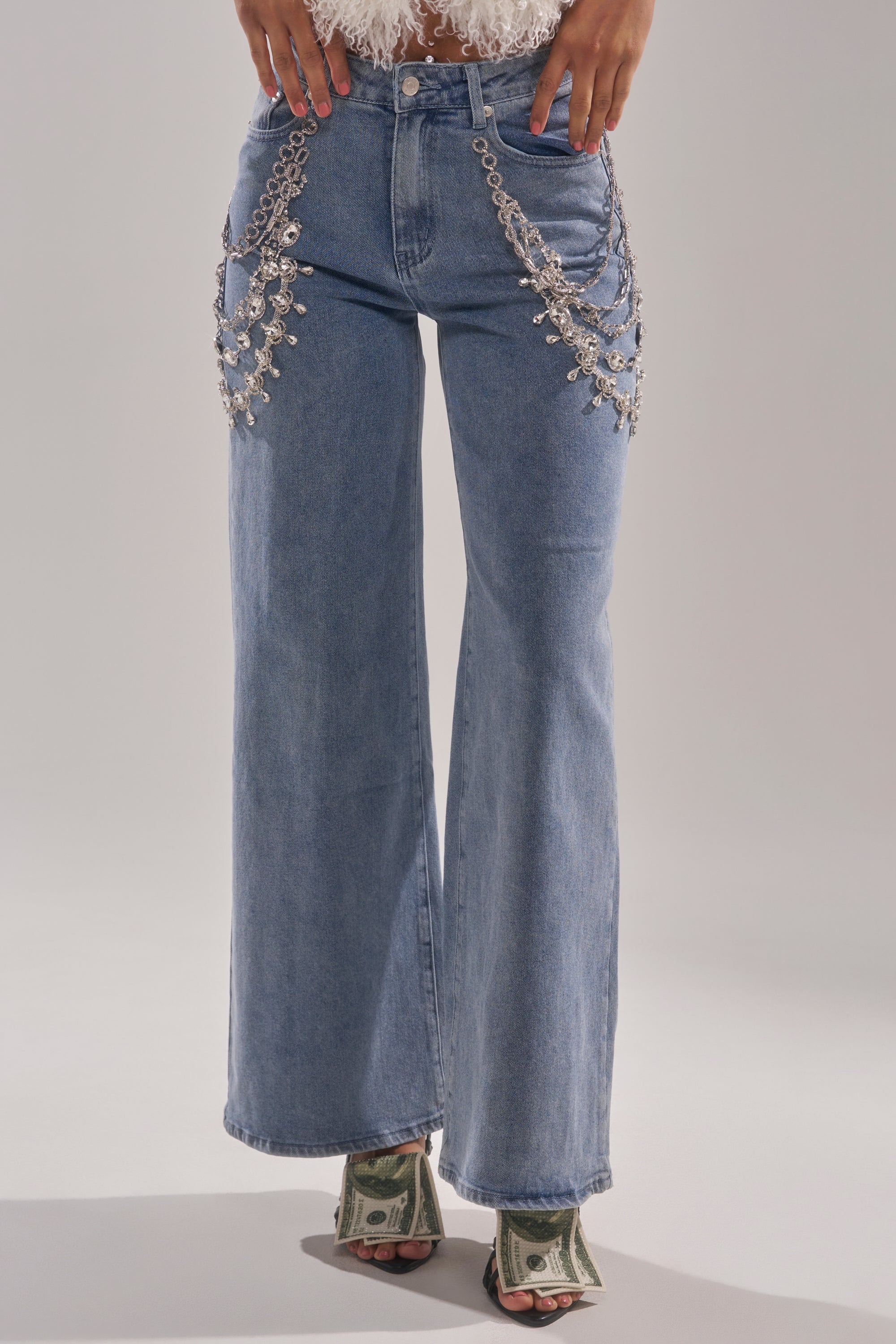 Wearing DIAMOND LOVER RELAXED DENIM WITH RHINESTONE—high-waisted, wide-leg blue jeans with rhinestones and silver chains—a person stands in open-toe heels wrapped in dollar bills against a plain background, showing only the lower body. 1