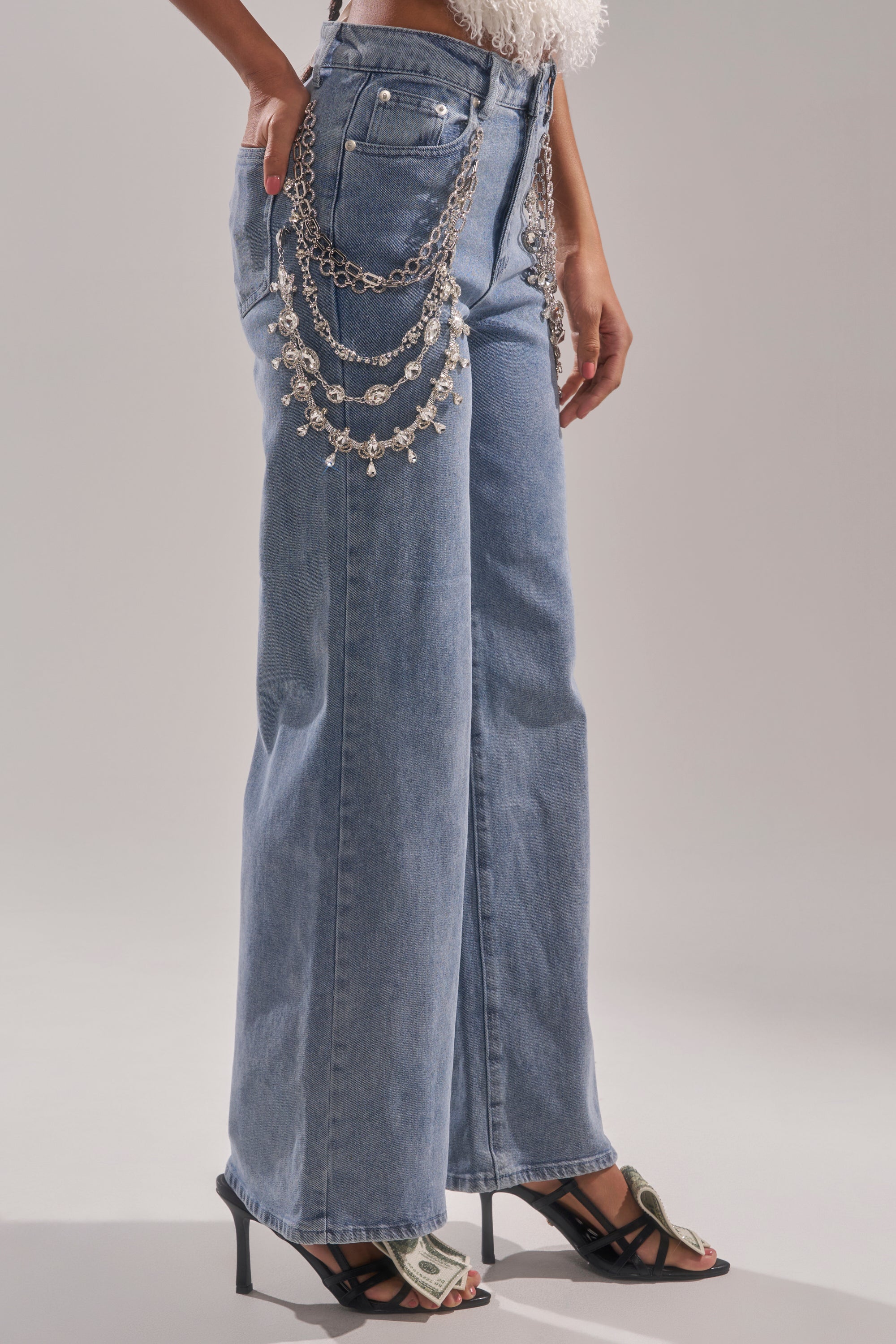 A person wears DIAMOND LOVER RELAXED DENIM WITH RHINESTONE, high-waisted wide-leg jeans, styled with a silver chain belt and black high-heeled sandals decorated with dollar bills, standing against a plain background. 2