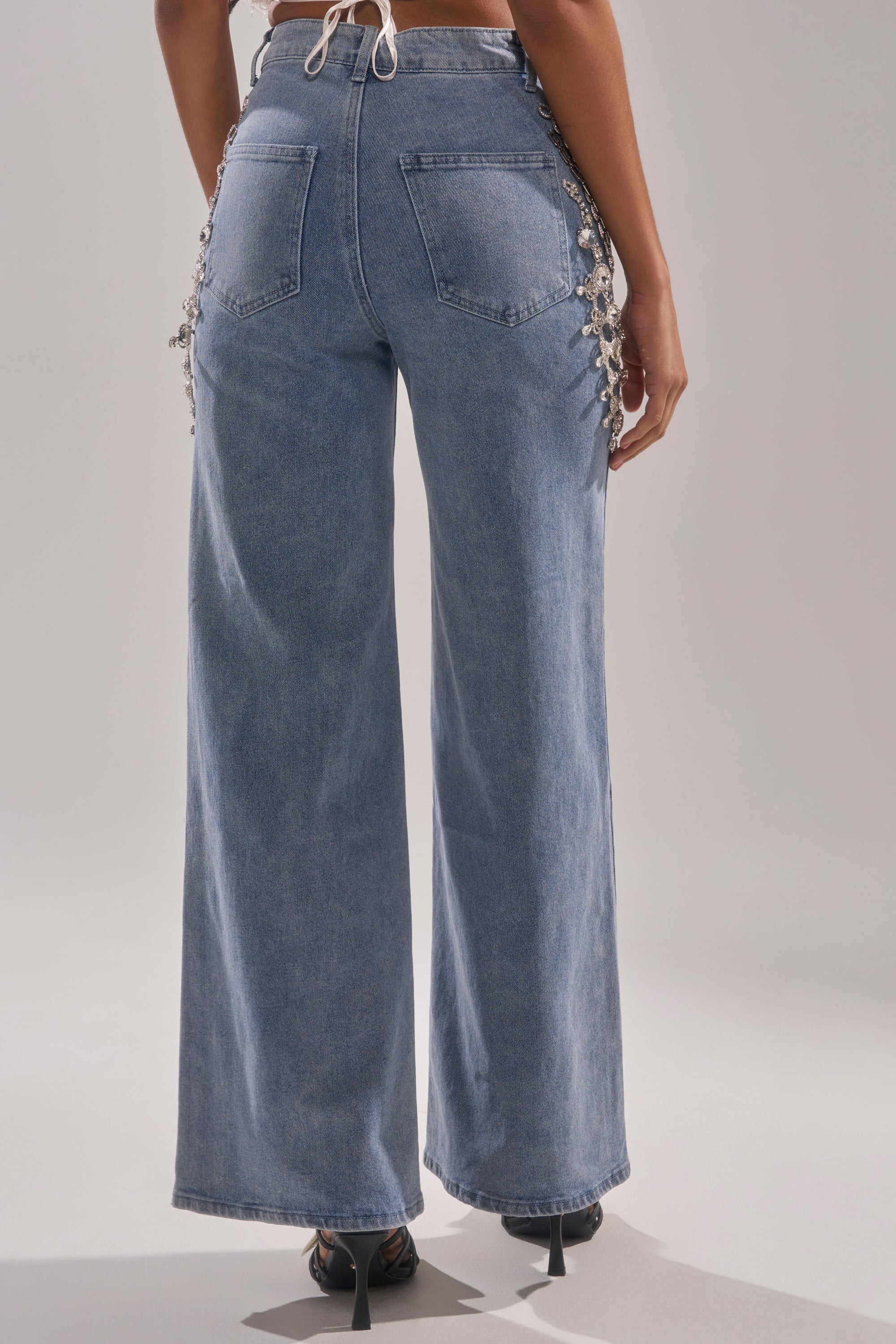 A person is seen from the back wearing DIAMOND LOVER RELAXED DENIM WITH RHINESTONE, featuring wide-leg blue jeans with decorative lace-up side details, standing against a plain light background. 3