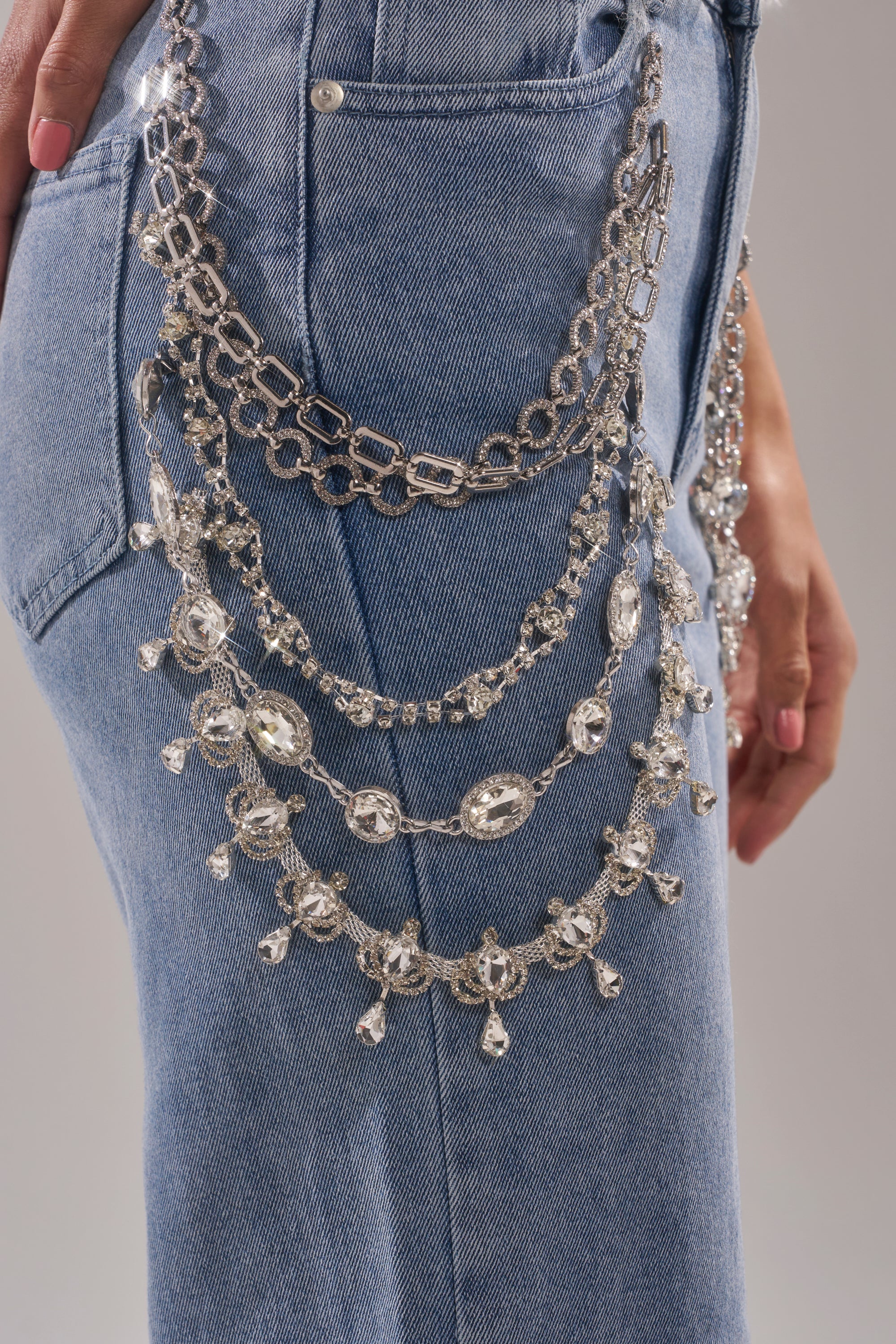 Wearing DIAMOND LOVER RELAXED DENIM WITH RHINESTONE, a person showcases relaxed wide-leg jeans featuring three layered ornate silver chains from the waistband to pocket, their hand resting by their side. 7