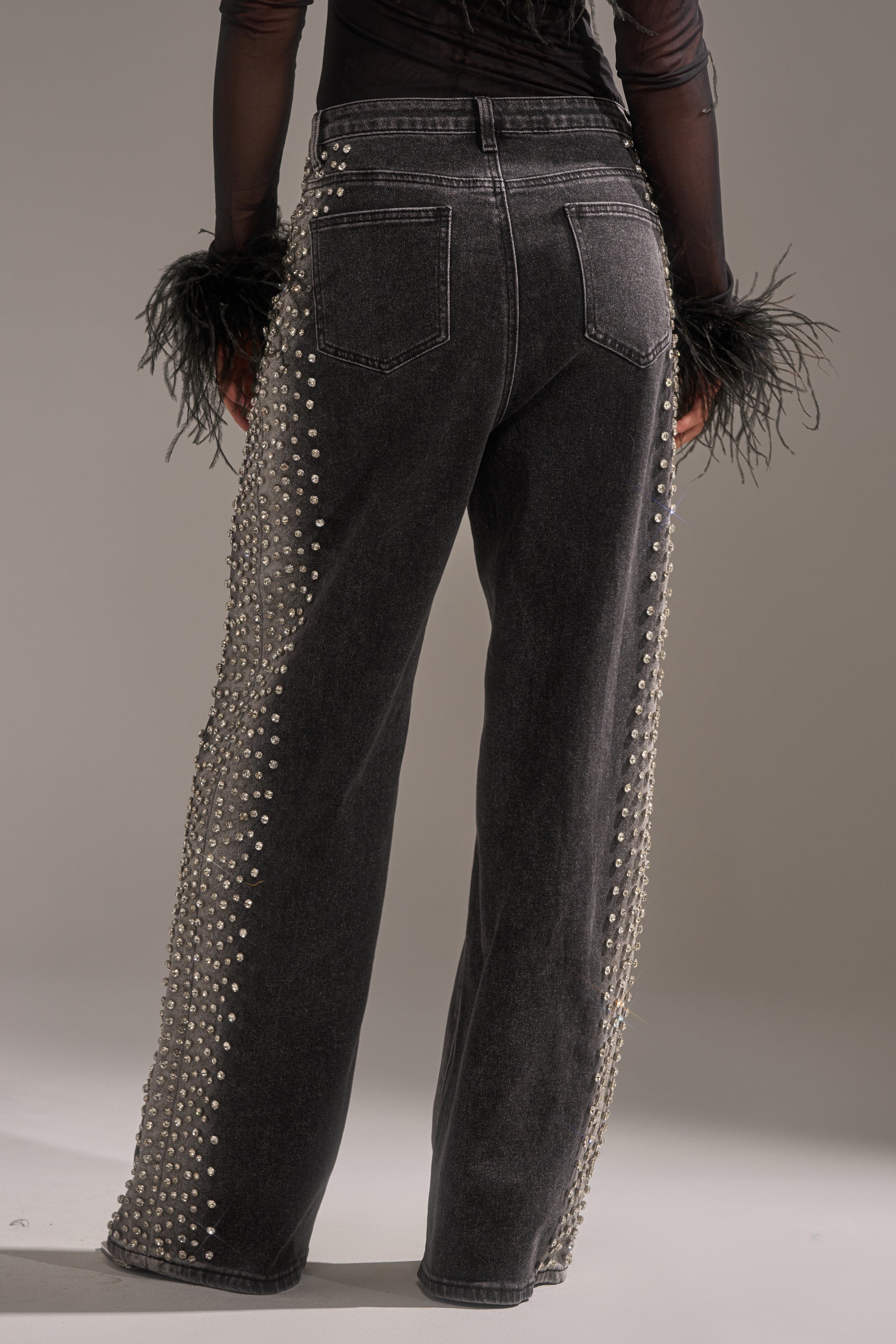 A person wears FREAKING FIRE EMBELLISHED DENIM—black wide-leg jeans adorned with pearl rows along the side seams—paired with a sheer black top with feathered cuffs. The image highlights the back view from waist down.