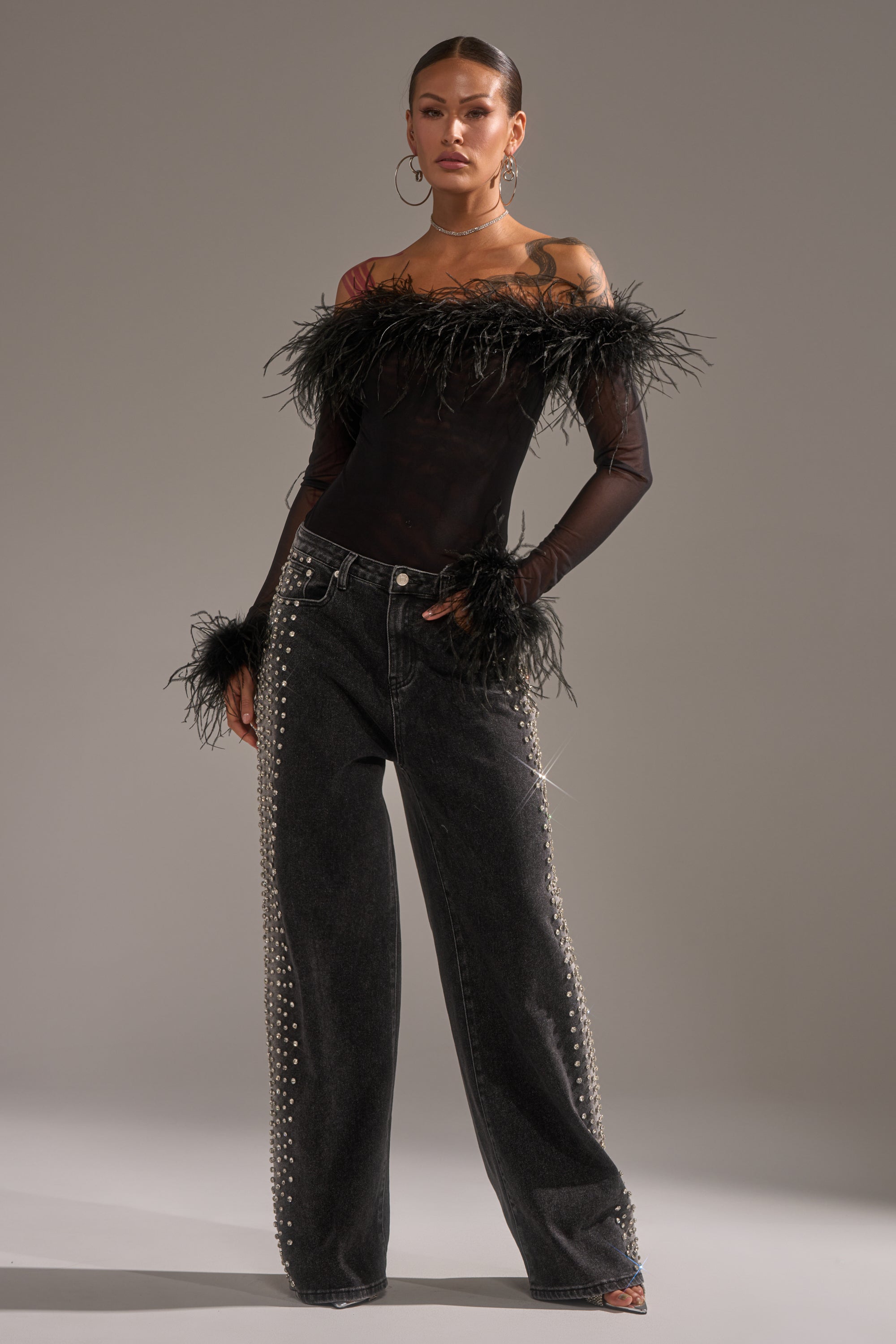 A woman poses confidently in a sheer black off-shoulder top with feather details, paired with FREAKING FIRE EMBELLISHED DENIM featuring loose legs and rows of metallic studs down the sides for a bold statement look.