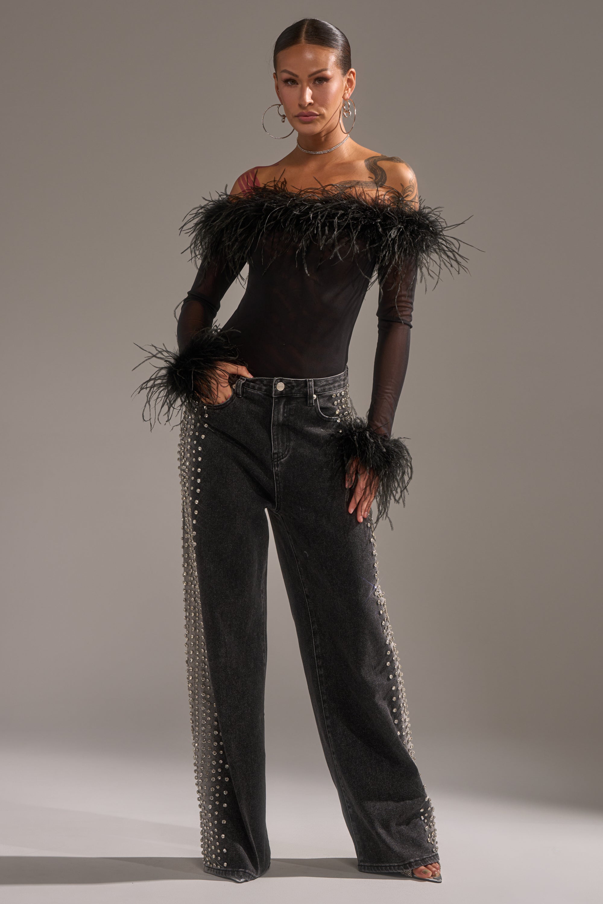 A woman stands confidently in an off-the-shoulder black top with feather trim and sheer sleeves, paired with the FREAKING FIRE EMBELLISHED DENIM—high-waisted, wide-leg jeans detailed with rows of silver studs along the sides.