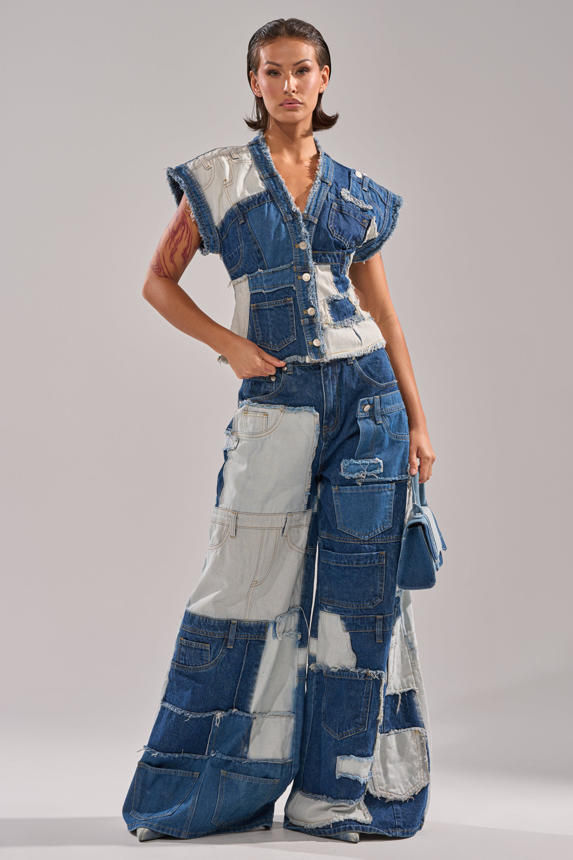 A person stands confidently in the TOO MUCH GOING ON WIDE LEG DENIM, a patchwork of various denim shades, paired with a matching bag and slicked-back hair for a bold, modern style.