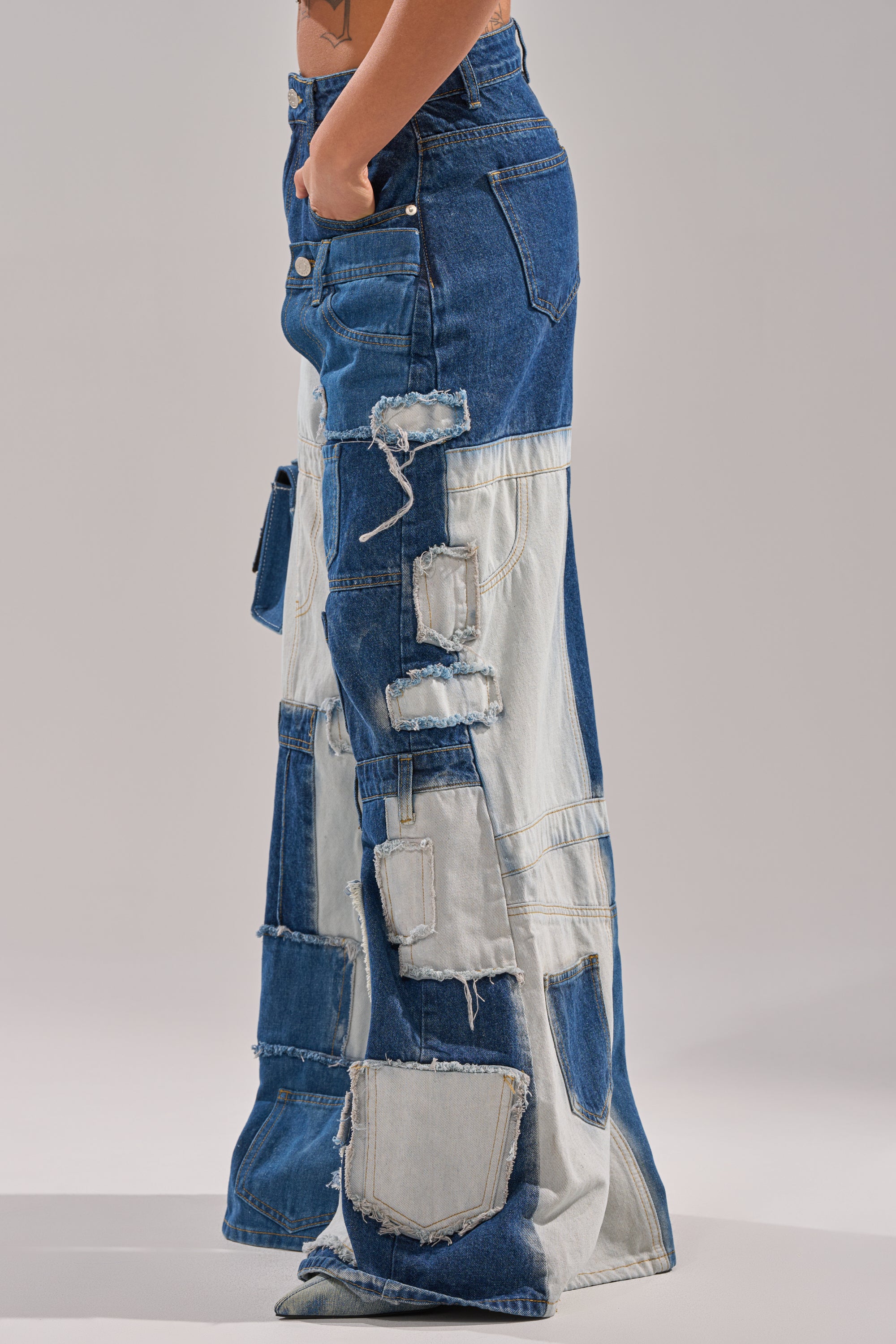 A person models the TOO MUCH GOING ON WIDE LEG DENIM—high-waisted, wide leg patchwork jeans in blue hues with frayed edges and distressed details—standing against a plain background, one hand in their pocket. 4