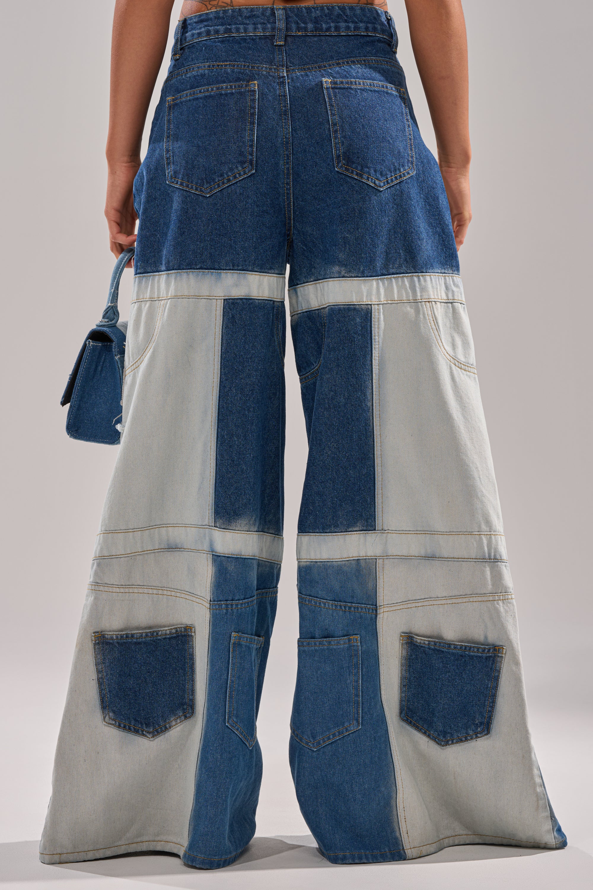 A person stands facing away, wearing the TOO MUCH GOING ON WIDE LEG DENIM—high waisted jeans with light and dark patchwork panels and multiple back pockets—while holding a small blue handbag. 5