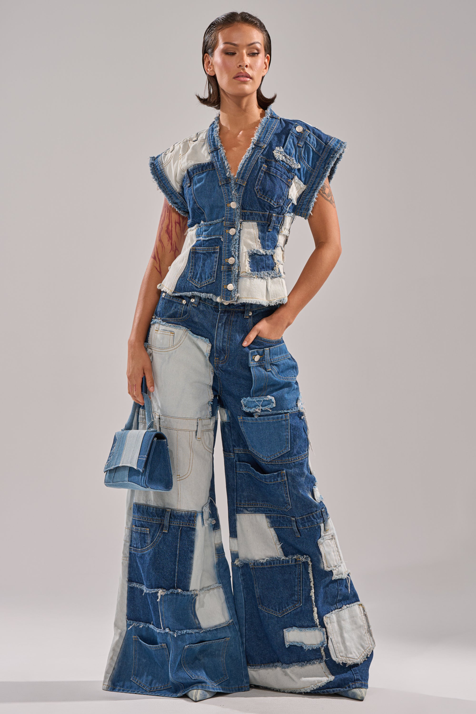 A woman stands confidently in the TOO MUCH GOING ON WIDE LEG DENIM, high waisted patchwork pants paired with a sleeveless button-up top and matching denim handbag, all featuring blue and white patchwork details. 6