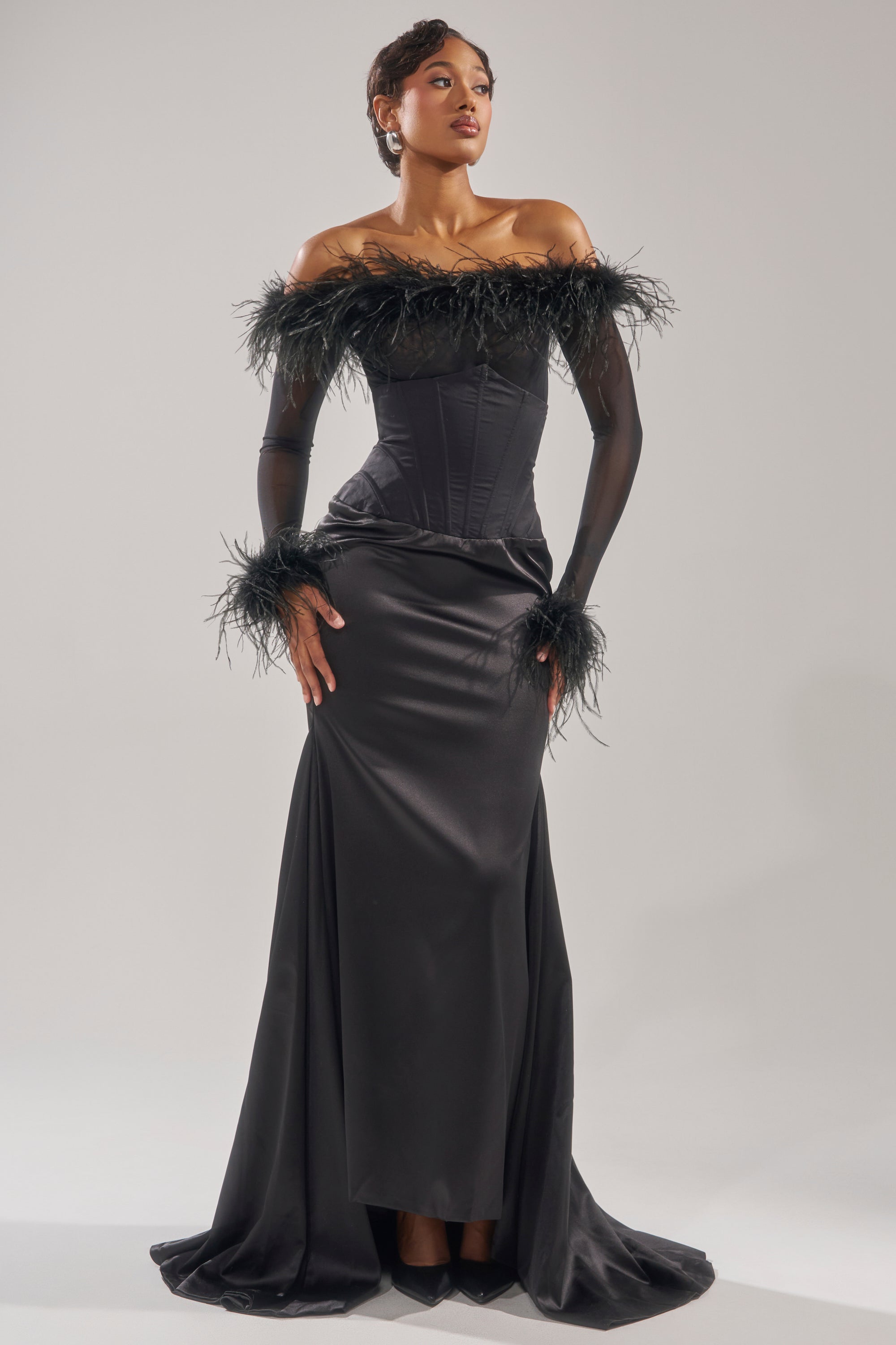 A woman poses in the STAR OF THE NIGHT HIGH WAISTED CORSET MAXI SKIRT—an elegant black maxi with feathered trim, corset waistband, and fitted bodice, flowing gracefully to the floor against a plain background. 5