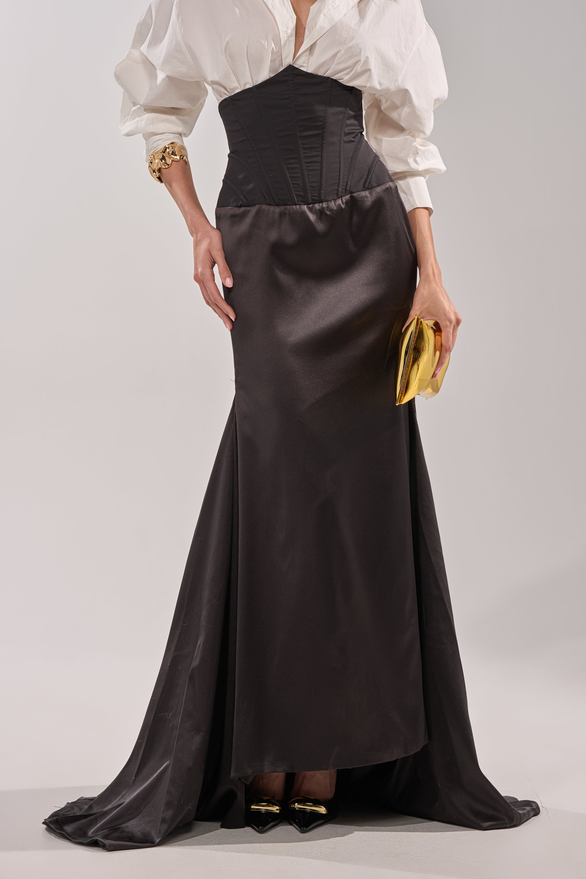 Wearing the STAR OF THE NIGHT HIGH WAISTED CORSET MAXI SKIRT in black satin, paired with a white blouse, gold clutch, and black pointed heels with gold accents. 1