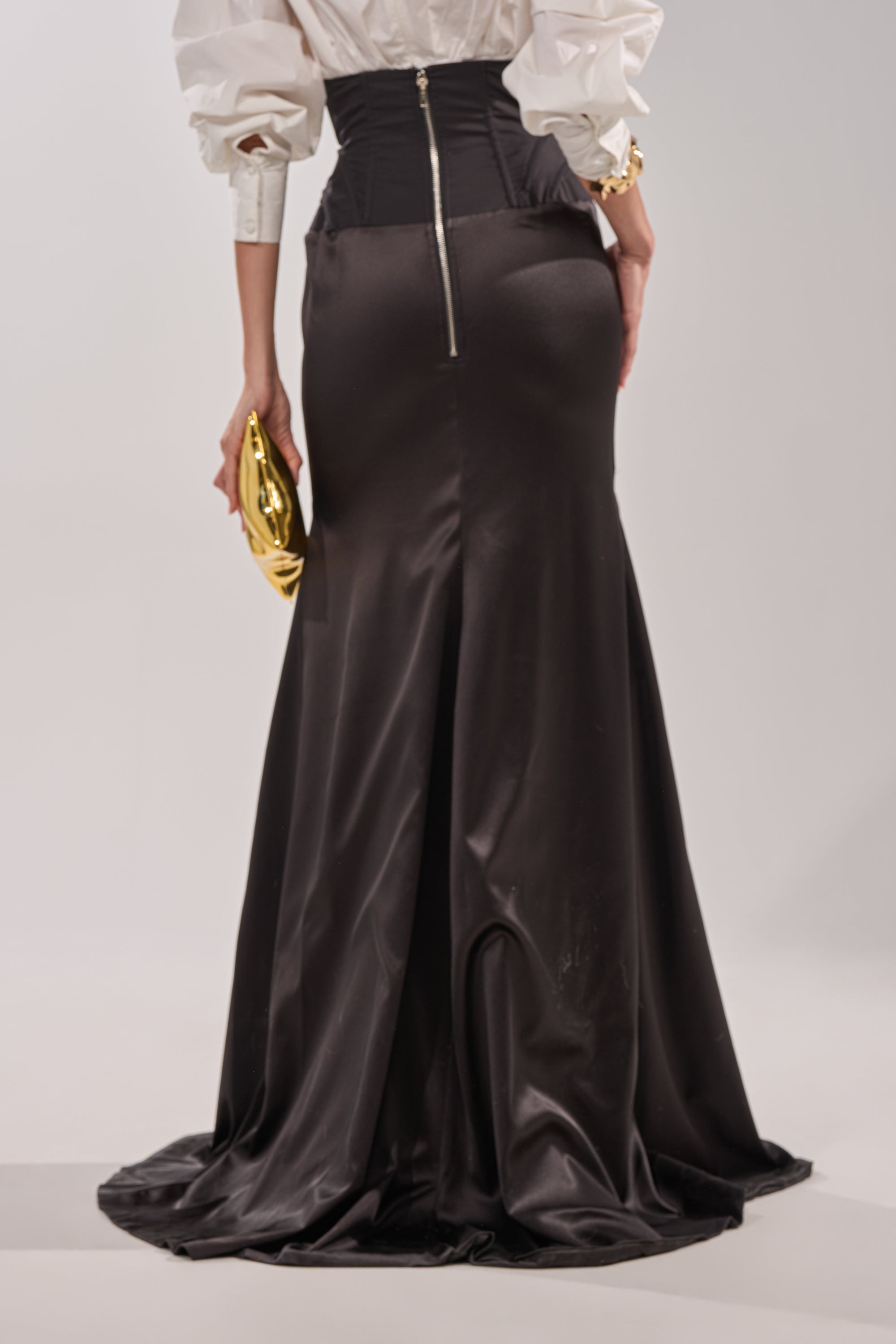A person is seen from behind in a STAR OF THE NIGHT HIGH WAISTED CORSET MAXI SKIRT, paired with a white shirt. They hold a shiny gold clutch and wear a gold bracelet, showcasing the skirt’s high waist and visible back zipper. 3