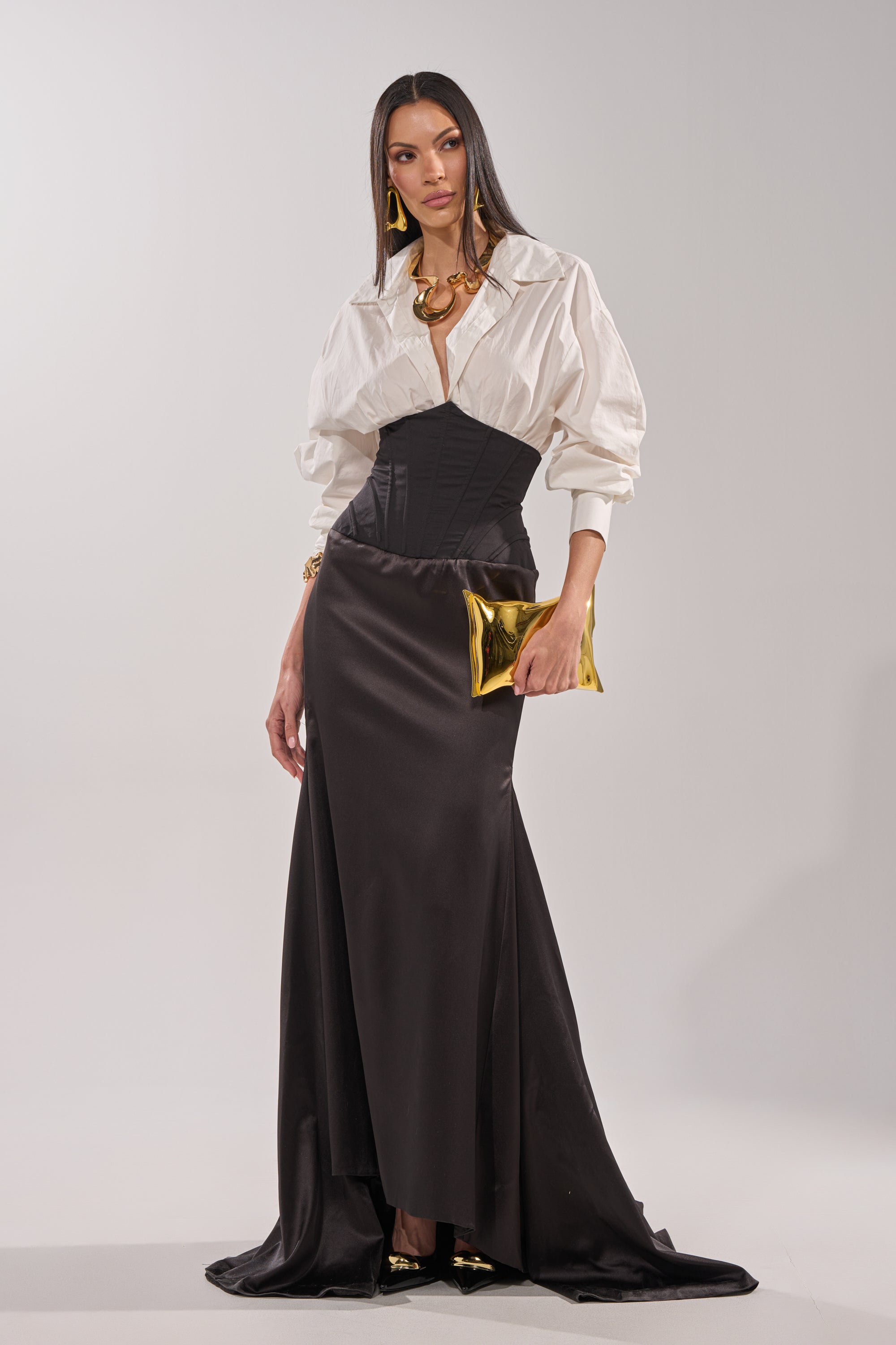 A woman poses against a plain background wearing the STAR OF THE NIGHT HIGH WAISTED CORSET MAXI SKIRT, styled with a white puff-sleeve blouse, black heels, bold gold jewelry, and a shiny gold clutch. 4