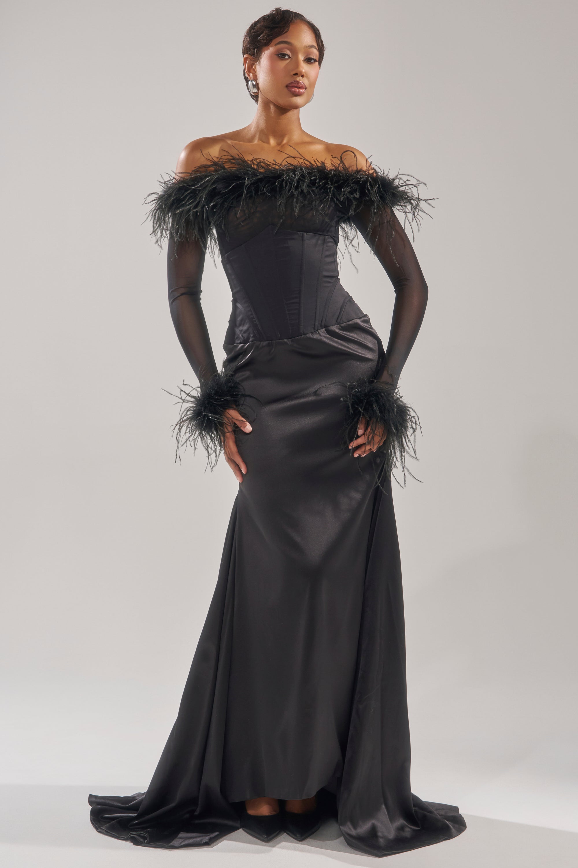 A woman poses gracefully in the STAR OF THE NIGHT HIGH WAISTED CORSET MAXI SKIRT, featuring a fitted corset waist and flowing satin mermaid silhouette with feather details, set against a light, simple background. 6