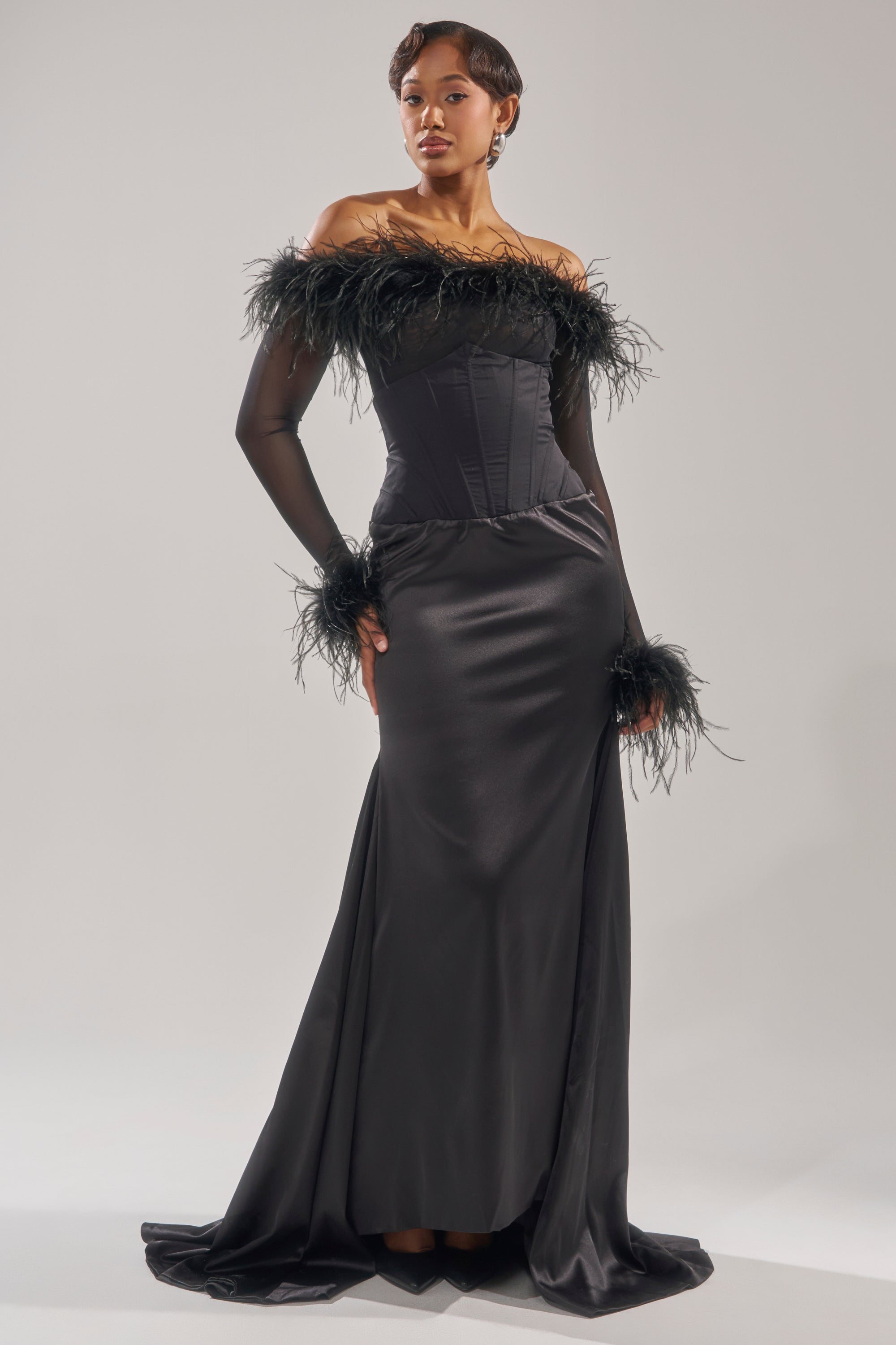 A woman poses against a plain light background, wearing the STAR OF THE NIGHT HIGH WAISTED CORSET MAXI SKIRT—a floor-length black skirt with a corset waistband, paired with an off-the-shoulder feather-trimmed sheer top. 7