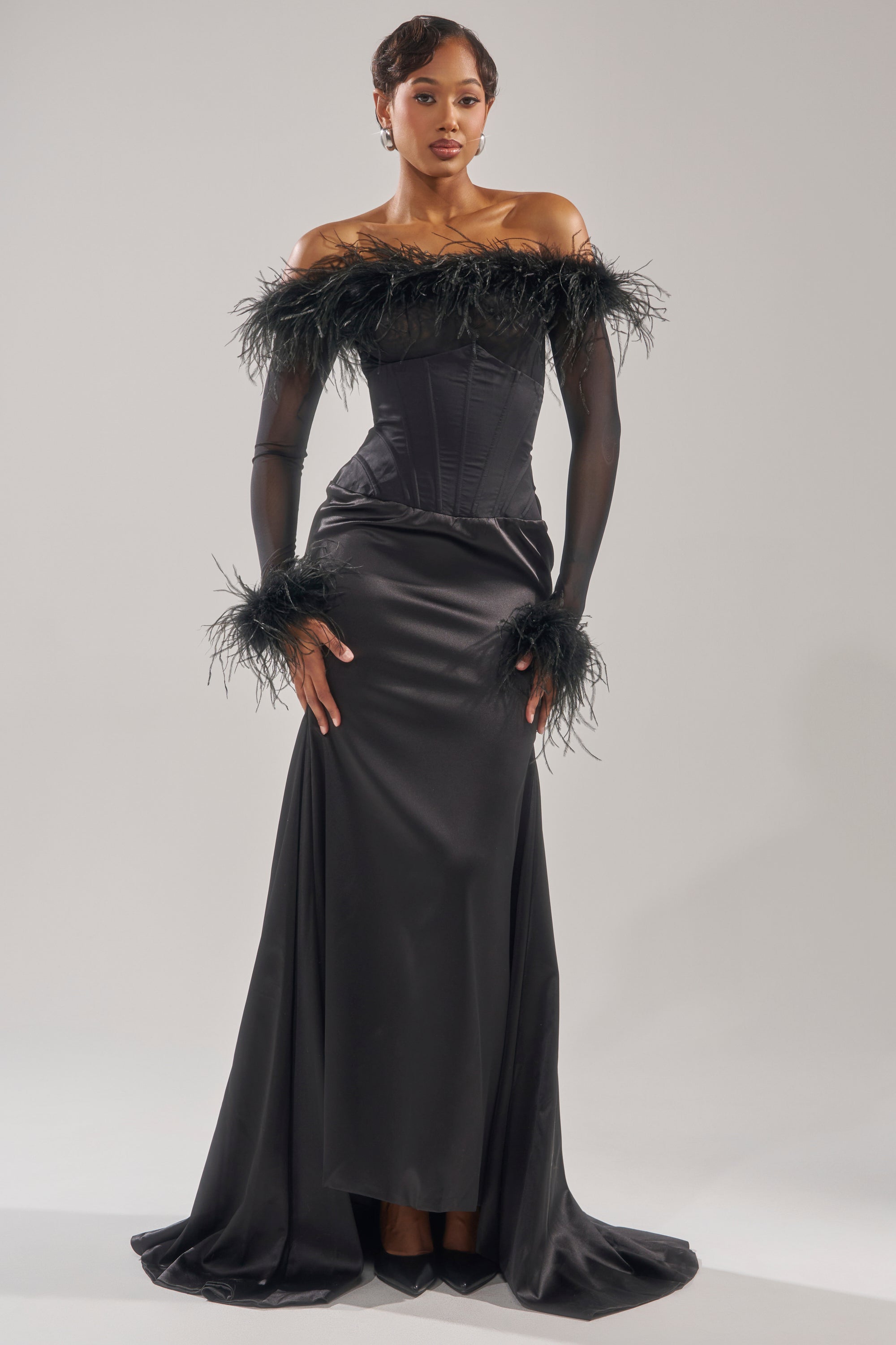A woman poses in the STAR OF THE NIGHT HIGH WAISTED CORSET MAXI SKIRT, a sleek black maxi with a corset waistband and sheer off-the-shoulder feather-trimmed sleeves, creating an elegant and glamorous look. 8
