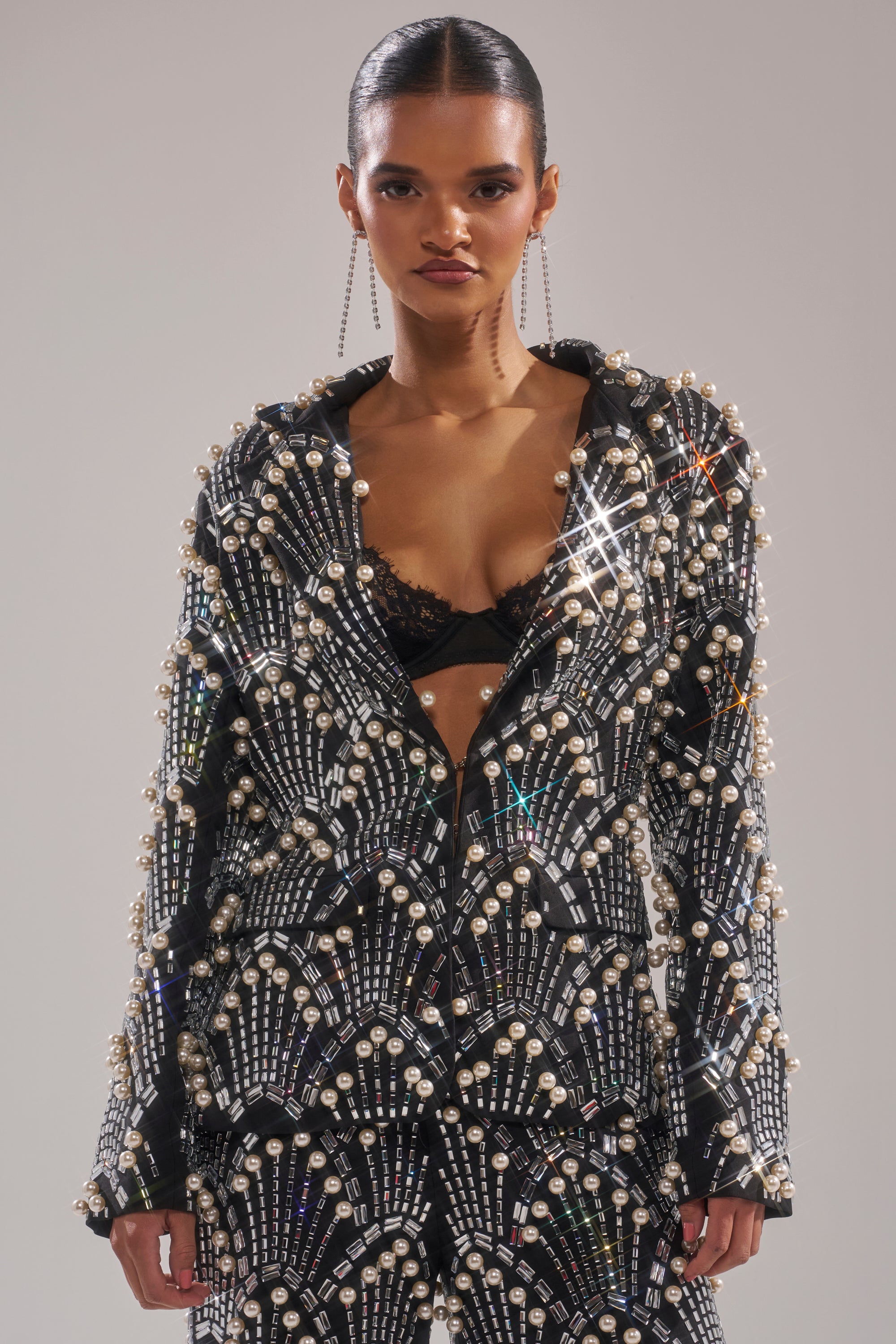 A model showcases the TIMES SQUARE EMBELLISHED BLAZER, detailed with pearls and crystals in geometric patterns. Worn open to reveal a black lace bra, the look is finished with slicked-back hair and dangling earrings.
