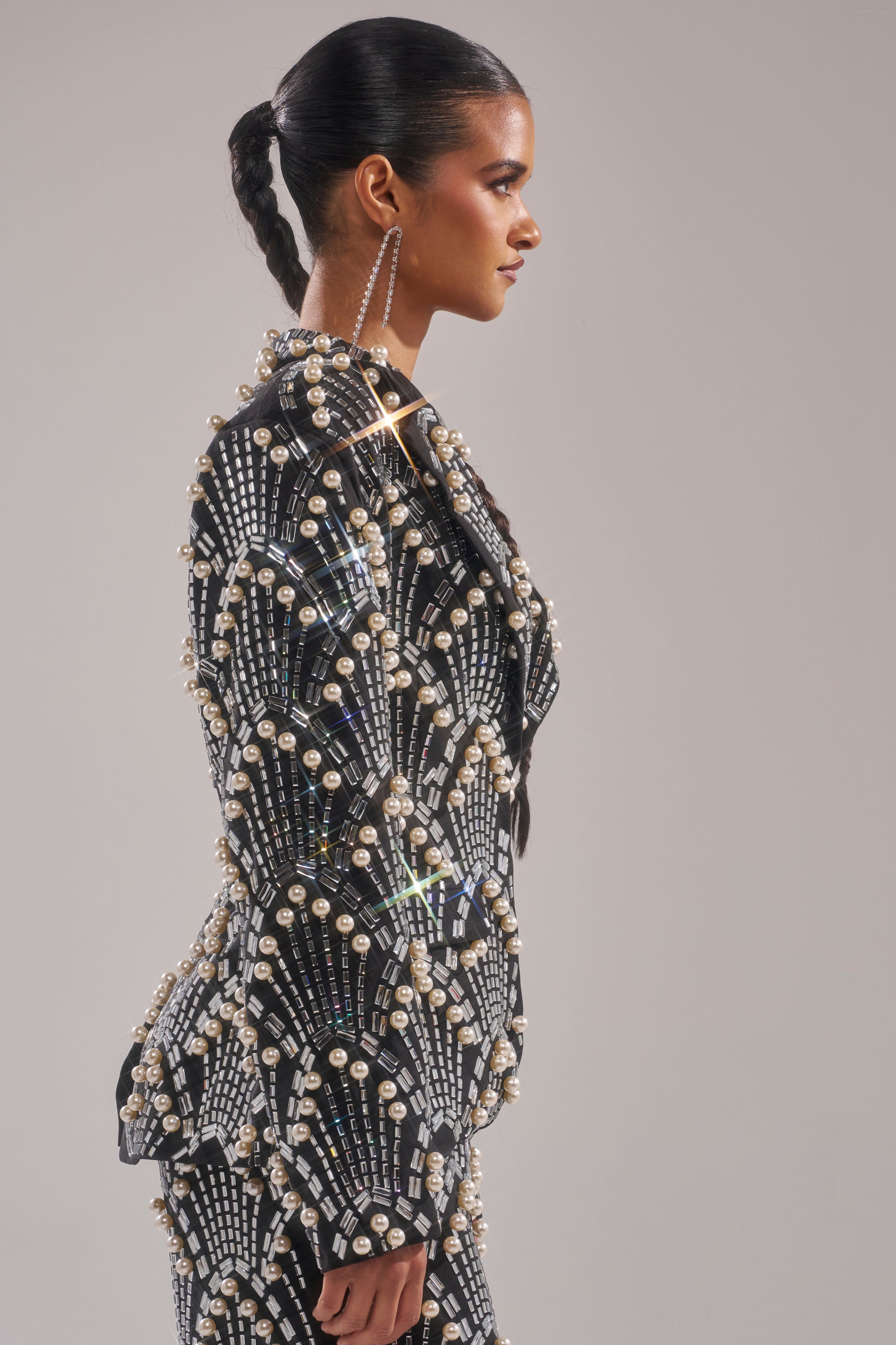 A woman with slicked back hair in a braid wears the fitted TIMES SQUARE EMBELLISHED BLAZER, featuring black fabric and sparkling pearl accents, shown in profile against a plain background.