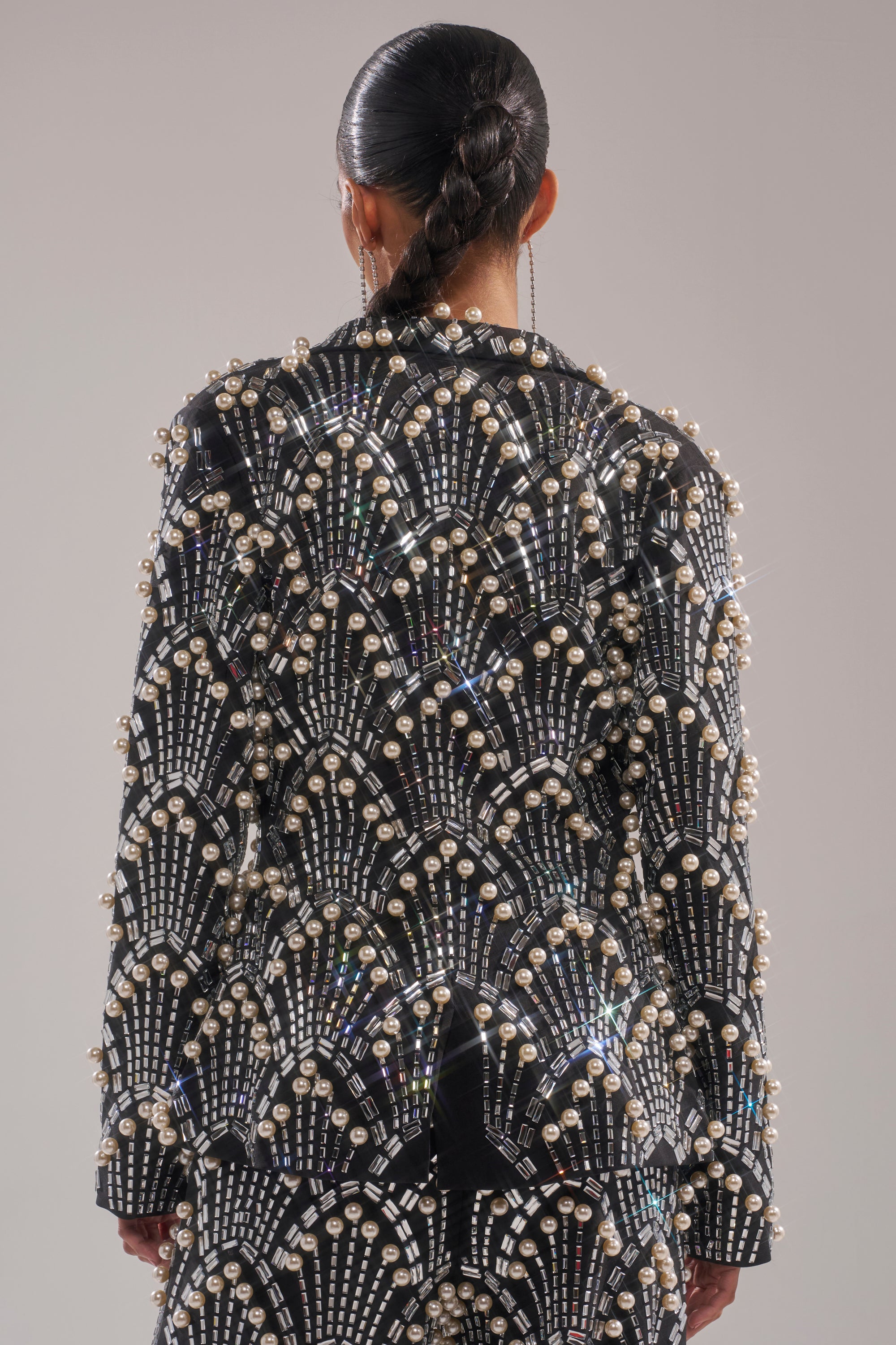 A person with slicked-back braided hair is seen from the back wearing the TIMES SQUARE EMBELLISHED BLAZER, featuring black fabric adorned with sparkling beads and pearls in geometric, fan-like patterns against a plain background.