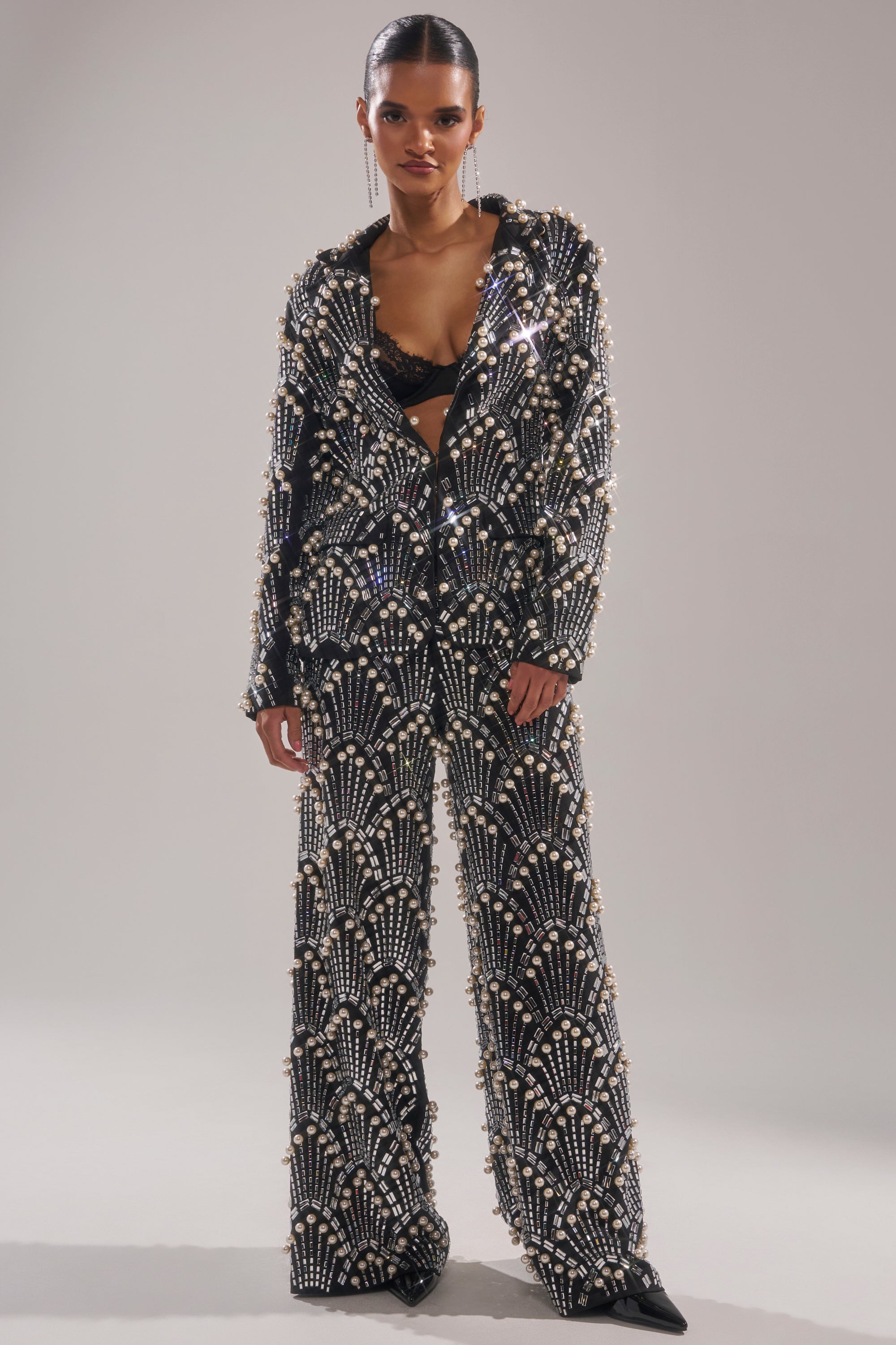 A person stands confidently in the TIMES SQUARE EMBELLISHED BLAZER, featuring white pearl and sequin patterns, paired with a black bralette and pointed shoes against a plain light background.