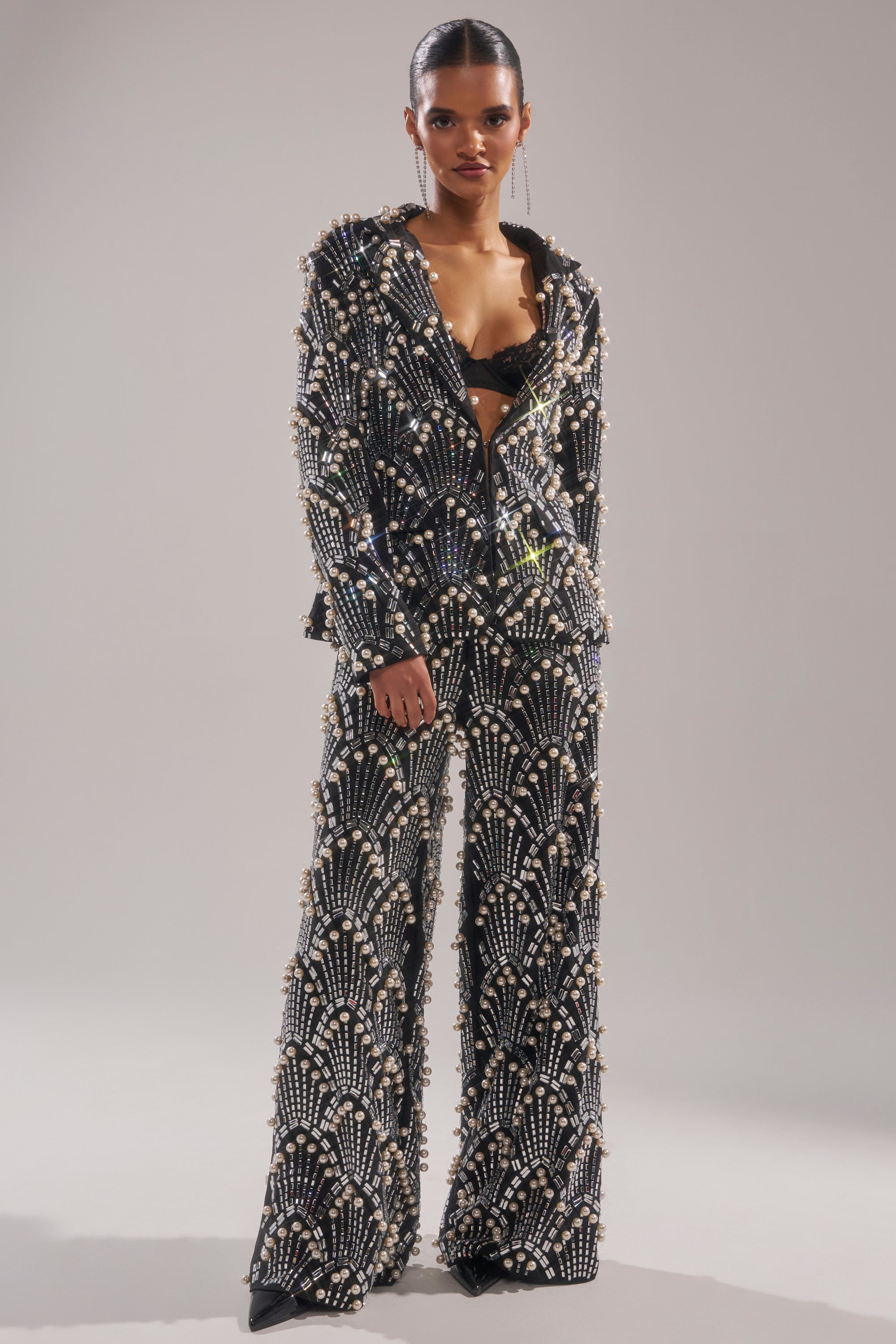A woman stands confidently in the TIMES SQUARE EMBELLISHED BLAZER, featuring elaborate silver and pearl beading, styled with a black bra top. Her sleek hair is pulled back as she poses against a plain backdrop with a neutral expression.
