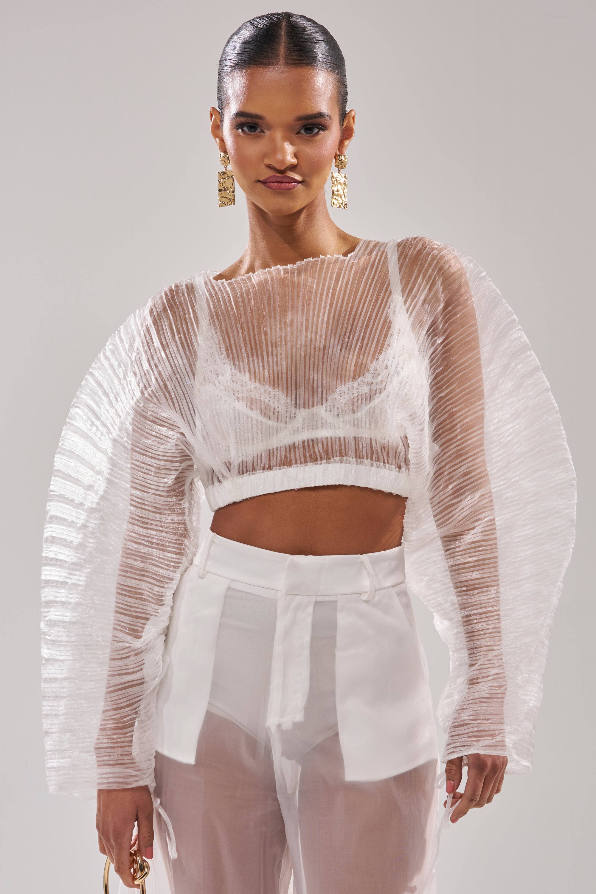 A woman models the VACATION MODE PLEATED BLOUSE—sheer and dramatic with curved sleeves—over a white bralette and high-waisted trousers, styled with gold earrings, a small bag, and slicked-back hair against a gray backdrop. 0