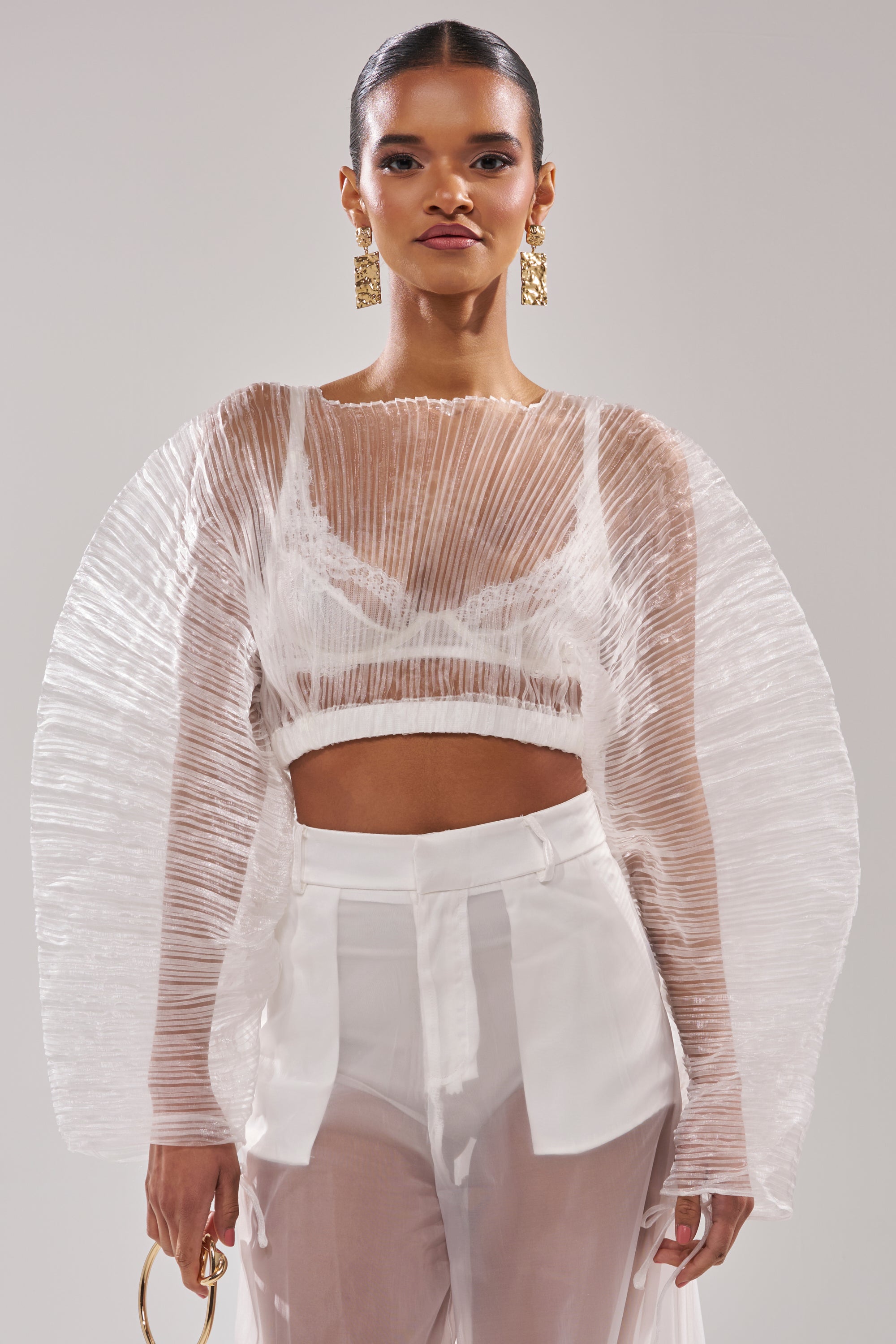 A woman models the VACATION MODE PLEATED BLOUSE—a sheer, resort-ready top with sculptural sleeves over a bralette, paired with semi-transparent high-waisted pants. She completes the look with bold gold earrings and a statement ring. 1