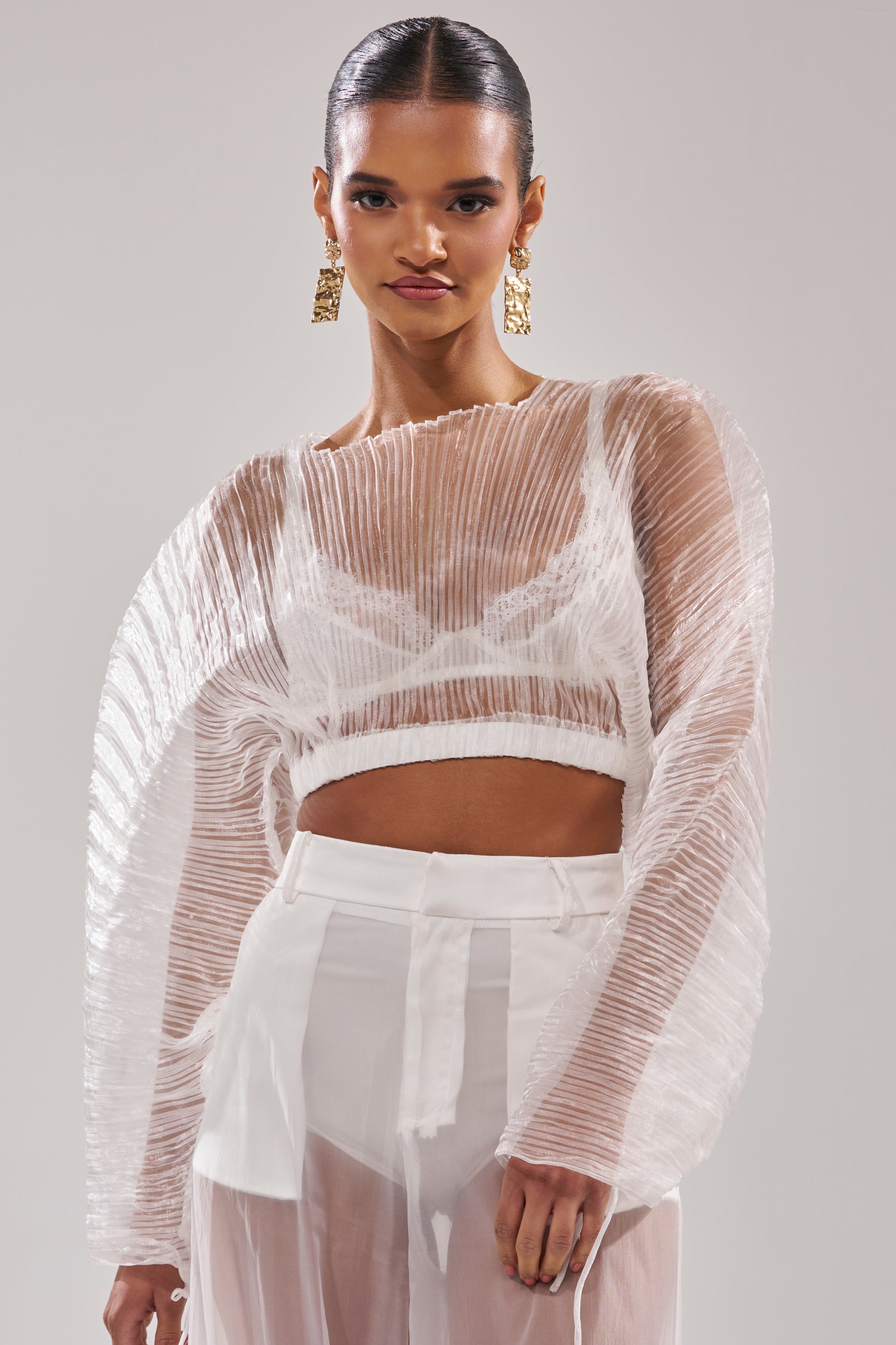 A woman in resort wear poses against a light background, styled in the VACATION MODE PLEATED BLOUSE with high-waisted sheer white pants, slicked-back hair, and bold gold earrings—ideal for your next getaway. 2