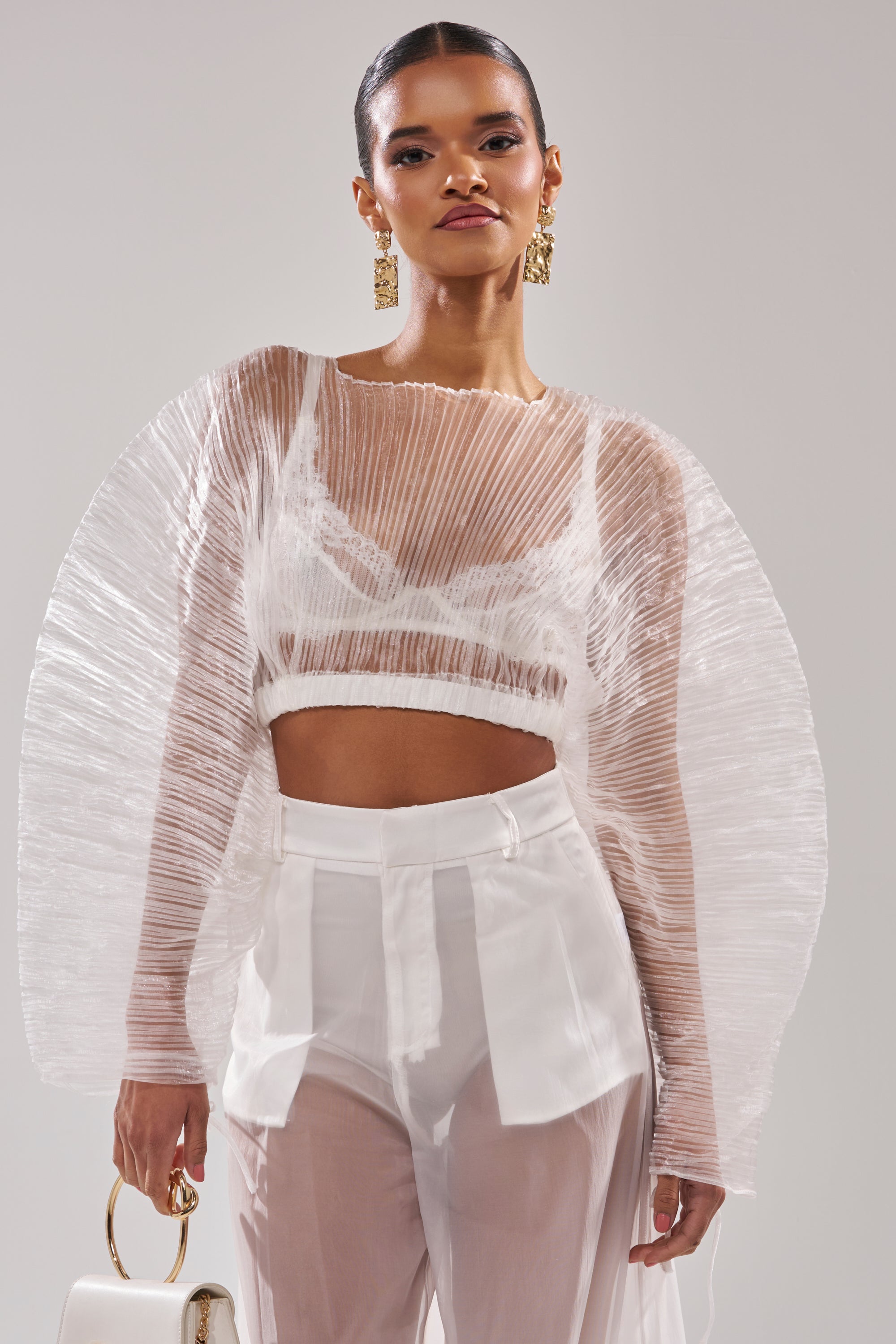 A woman models the VACATION MODE PLEATED BLOUSE—sheer and pleated with dramatic rounded shoulders—styled over a white bralette and high-waisted, semi-transparent pants, finished with gold earrings and a white handbag for chic resort wear. 3