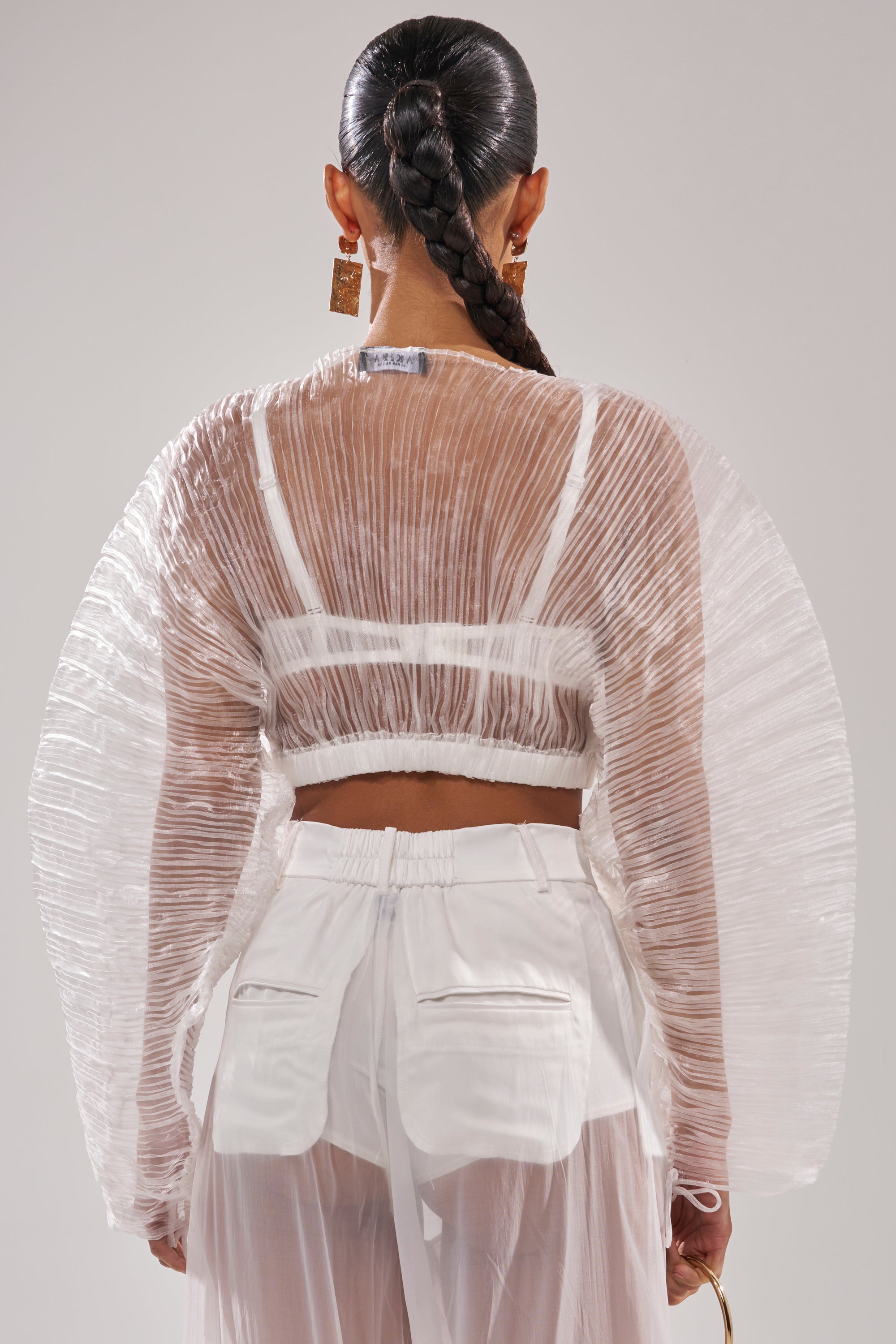 A person with a long braided ponytail models the VACATION MODE PLEATED BLOUSE, featuring dramatic wing-like sleeves, paired with sheer patch-pocket pants, posing with their back to the camera for an effortless resort style. 5
