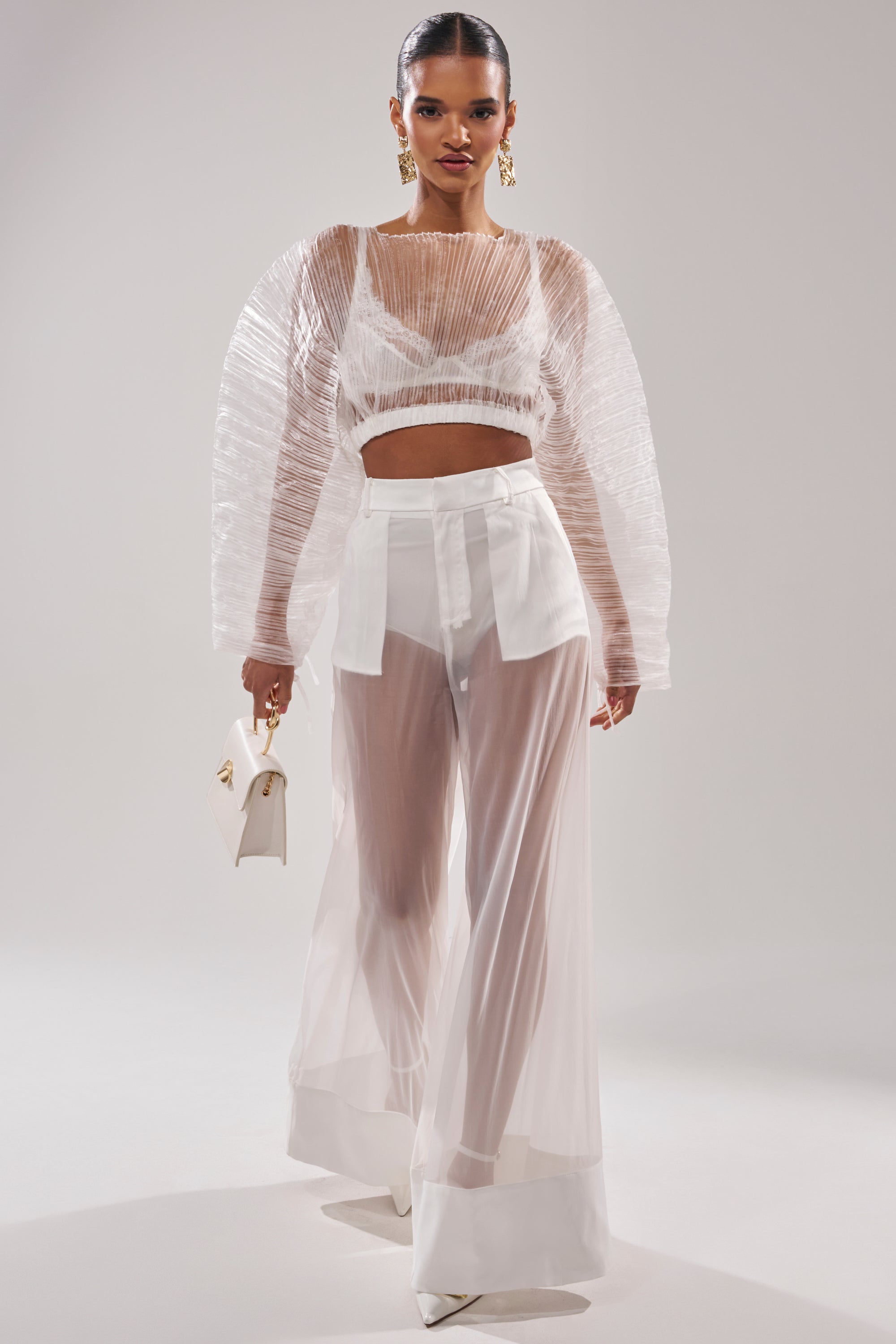 A woman models the VACATION MODE PLEATED BLOUSE, featuring airy pleats and voluminous sleeves, paired with wide-leg sheer white pants over shorts. She holds a white handbag and stands against a light background for a chic, resort-ready look. 6