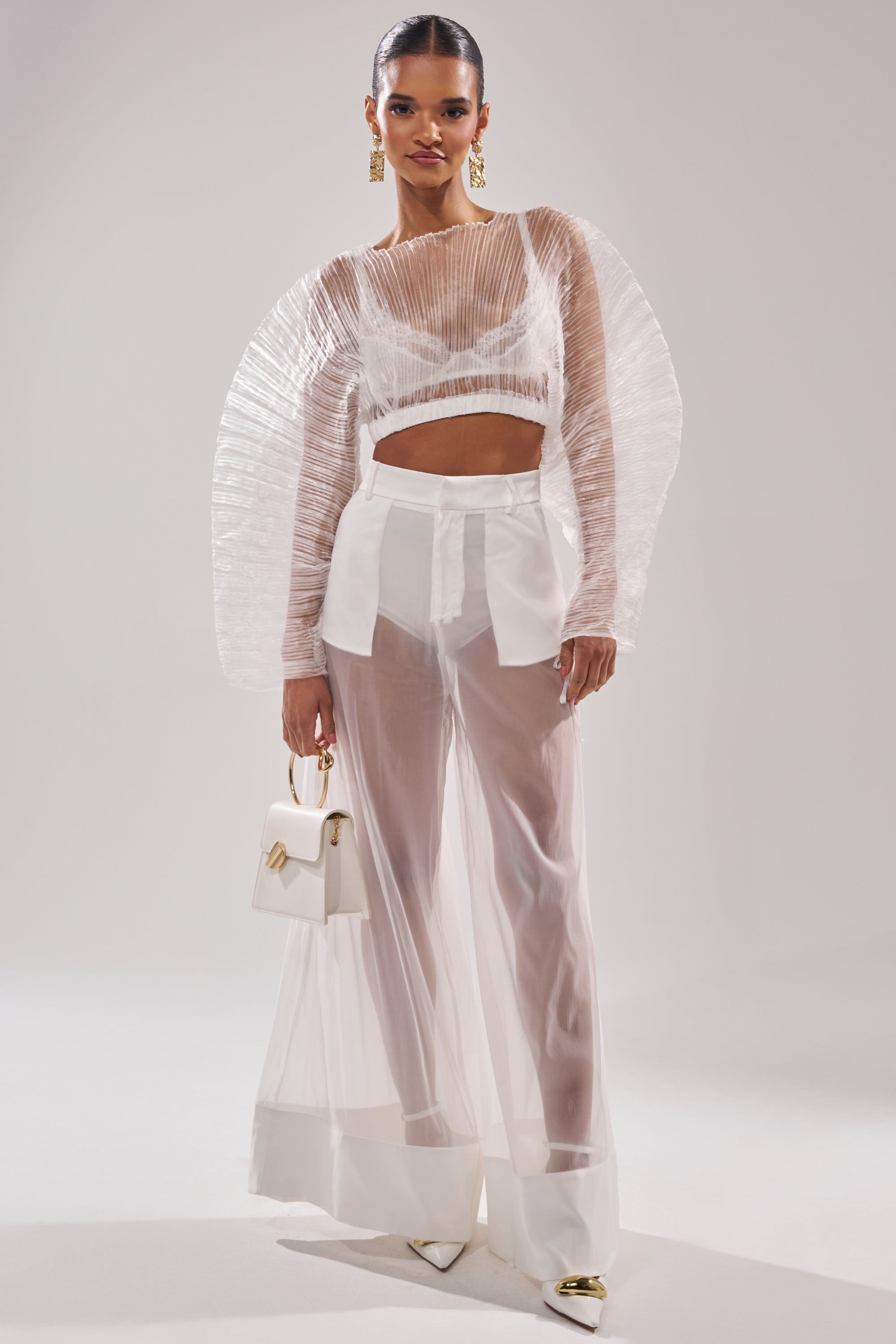 A woman models the VACATION MODE PLEATED BLOUSE in sheer white with wide-leg pants, accessorized with a small white handbag, white heels, and gold earrings, against a plain backdrop. 7