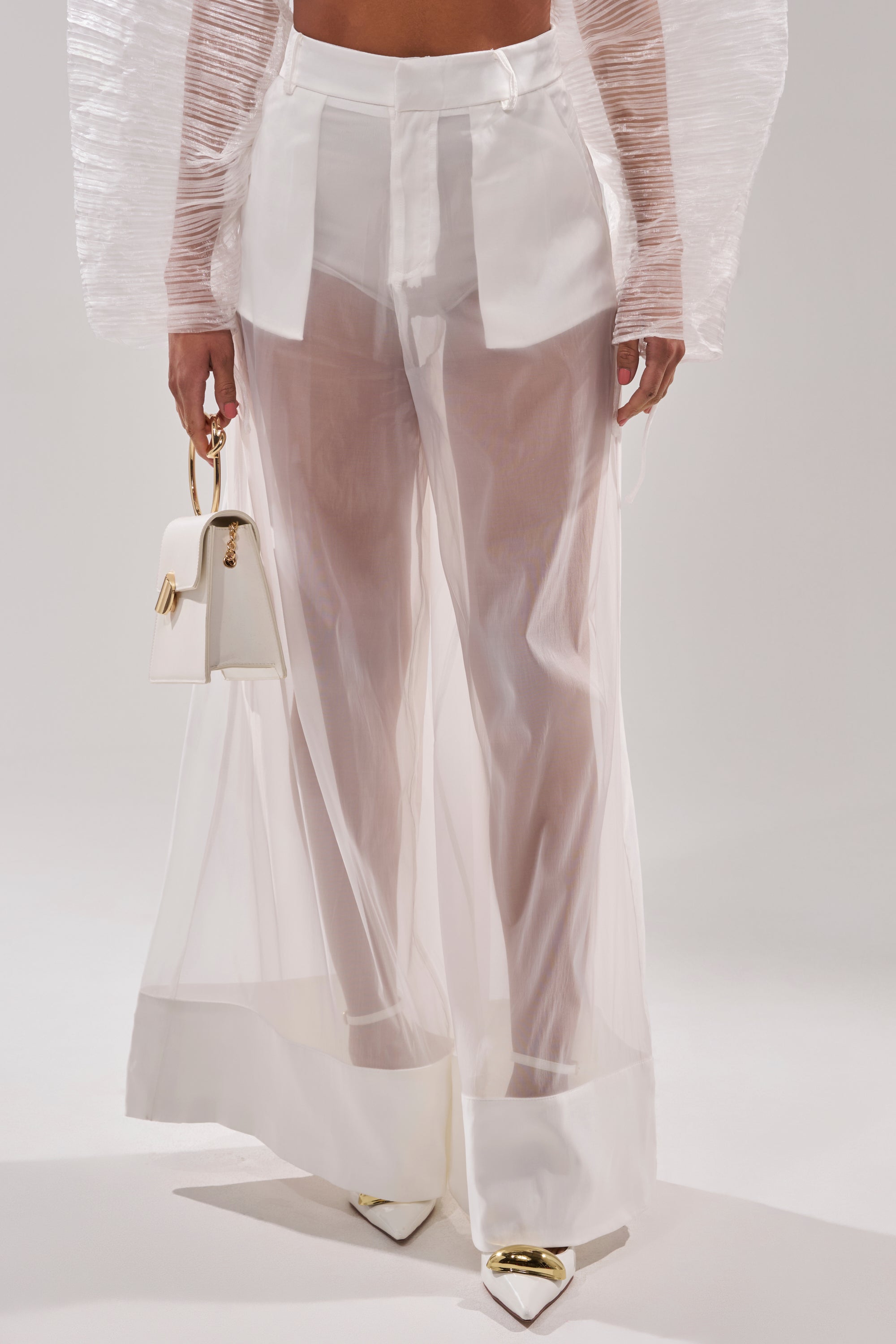 A person wearing the VACATION MODE TROUSER—high-waisted, sheer organza pants with solid white cuffs and waistband—paired with a translucent long-sleeve top, white pointed heels, and a small white handbag with gold hardware. 1