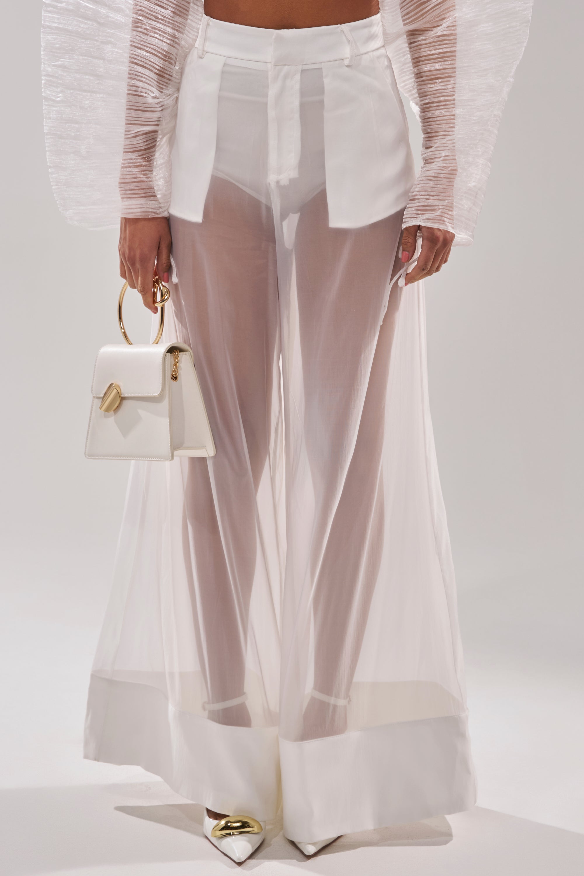 Wearing the VACATION MODE TROUSER, sheer organza wide leg pants over white shorts, paired with a white textured long-sleeve top, pointed white shoes with gold accents, and holding a small white handbag with a gold ring handle. 2