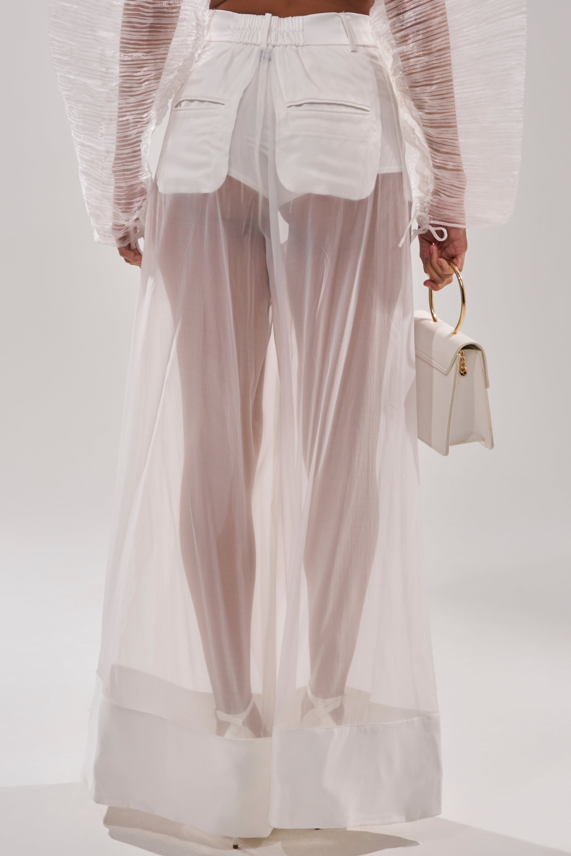 Wearing the VACATION MODE TROUSER—sheer organza pants layered over visible shorts—with a long-sleeve top featuring textured sleeves, and holding a small white handbag with a gold handle. The background is plain and light-colored. 4