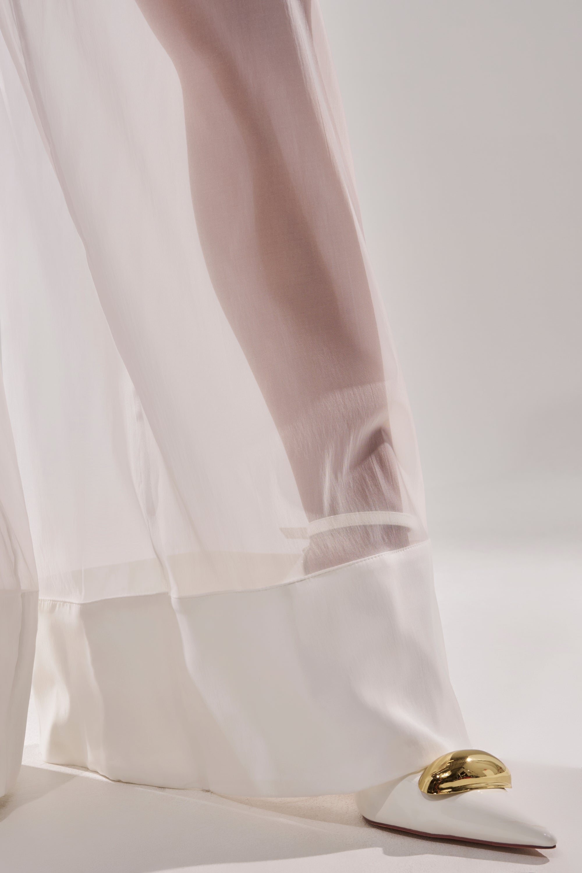 A close-up of the VACATION MODE TROUSER, a sheer white, floor-length style showing a leg beneath, paired with a white pointed shoe with a shiny gold toe accent against a neutral background. 6