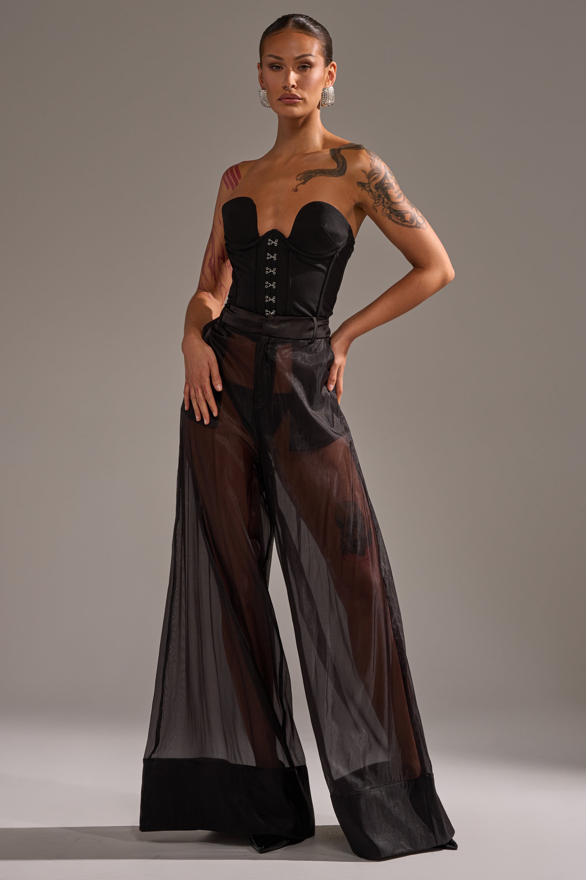 A woman poses confidently in the LOUD AND LUX TROUSER, paired with a black strapless corset top. Her tattoos accent her arm and shoulder, and her hair is pulled back. The outfit stands out against a plain gray background. 0