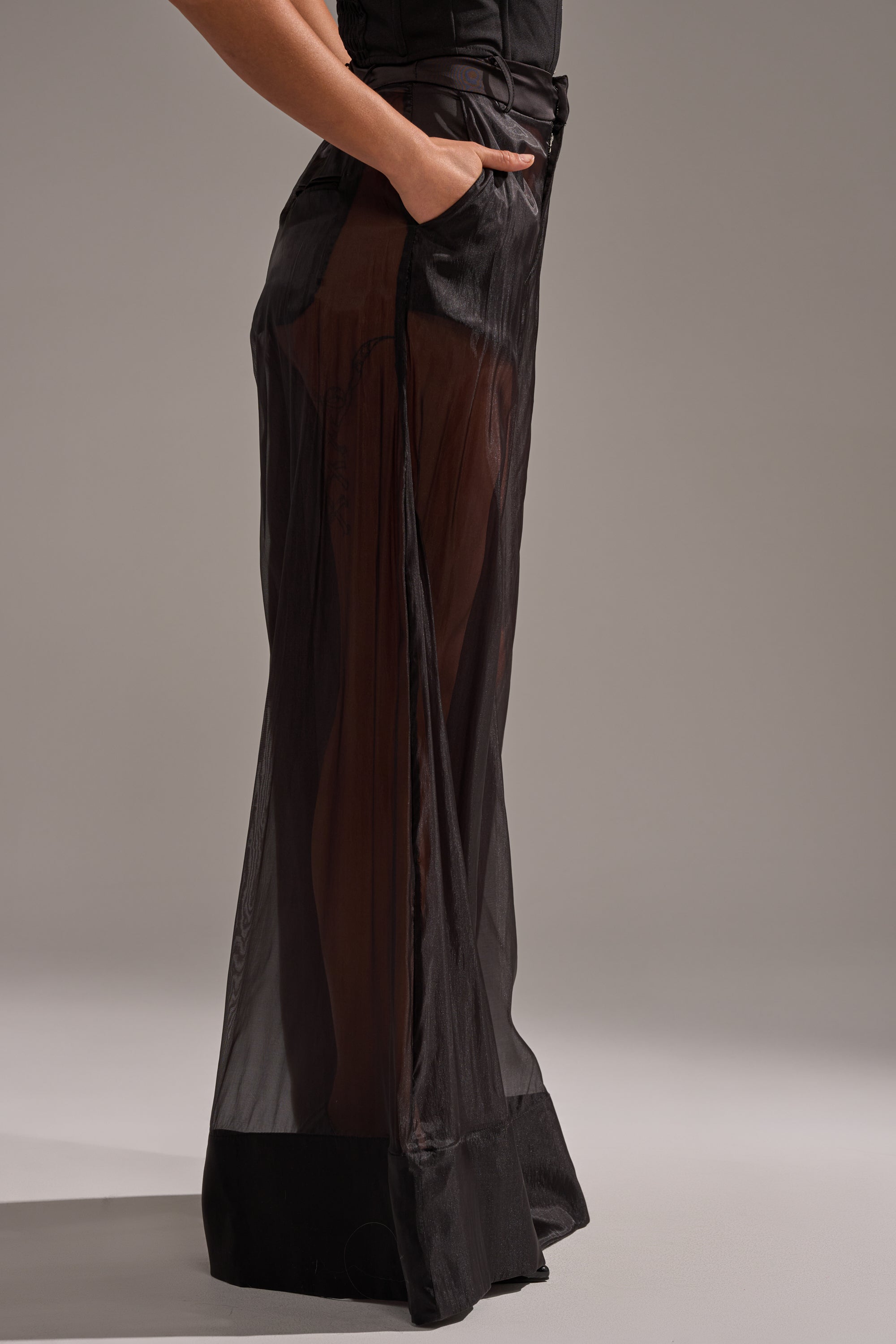 A person stands with one hand in their pocket, wearing the LOUD AND LUX TROUSER—sheer, black wide-leg pants that reveal their legs—against a plain, neutral background. 3