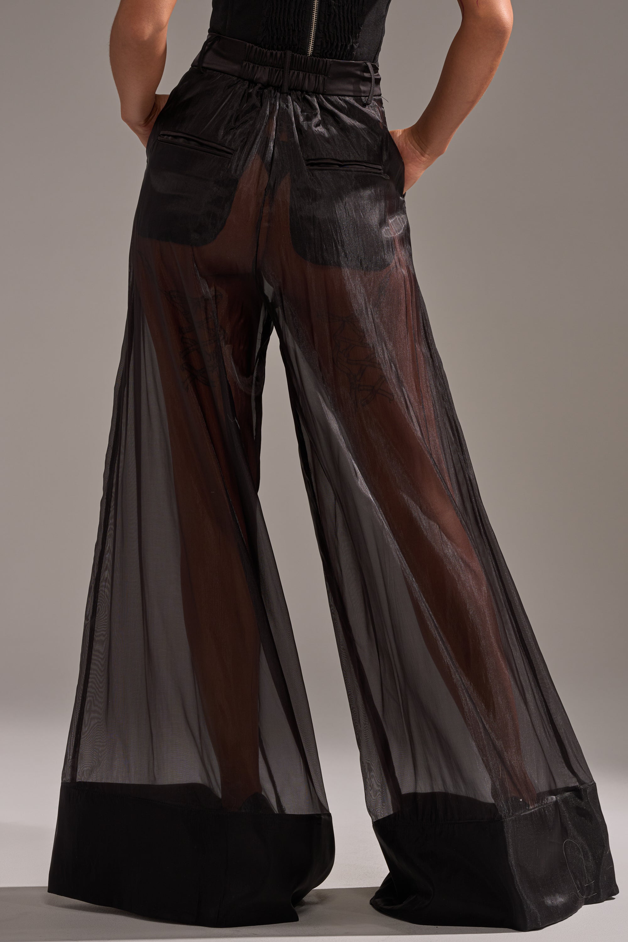 A person is standing against a plain background, wearing the LOUD AND LUX TROUSER—high-rise, wide-leg black sheer pants with solid black panels at the waistband, pockets, and cuffs that reveal their legs underneath. 4